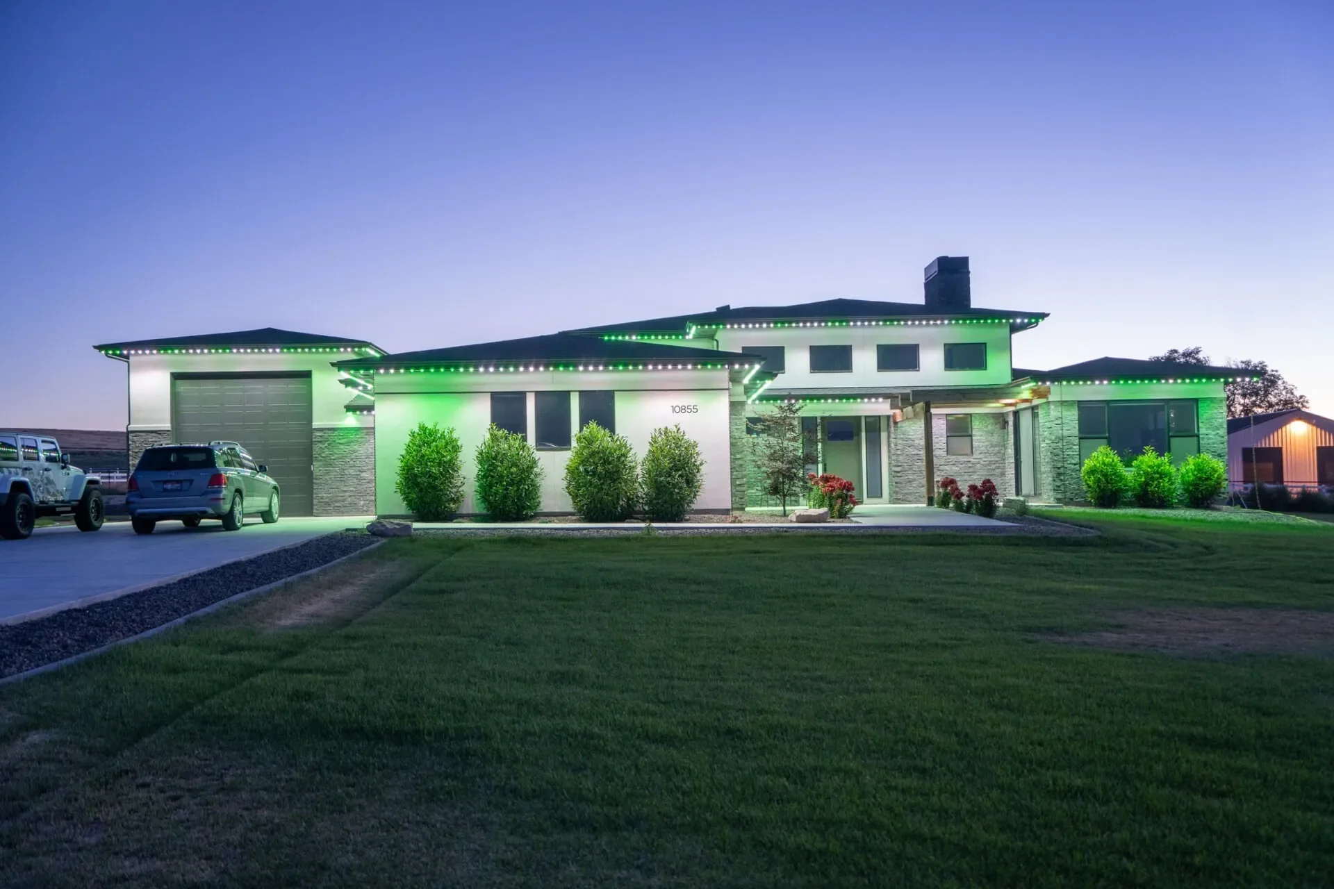 A modern house illuminated with green and white Christmas lights at dusk. The house has a large front lawn, a driveway with parked cars, and bushes lining the entrance.