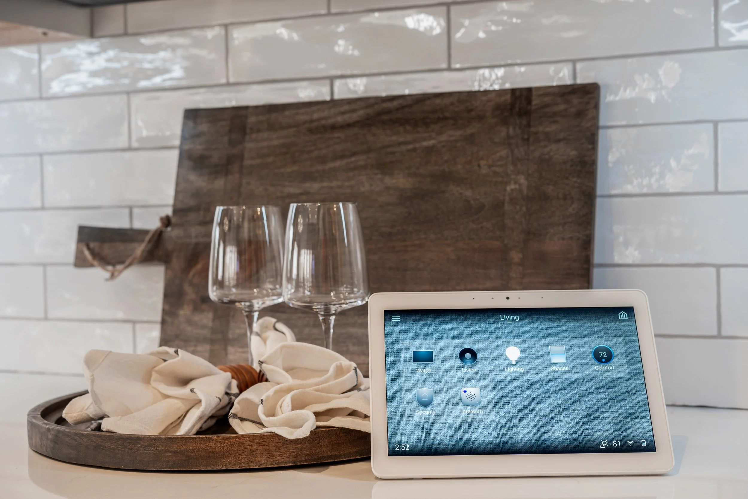 A smart home device on a kitchen counter with two empty wine glasses and a napkin on a wooden tray in the background against a white brick wall.