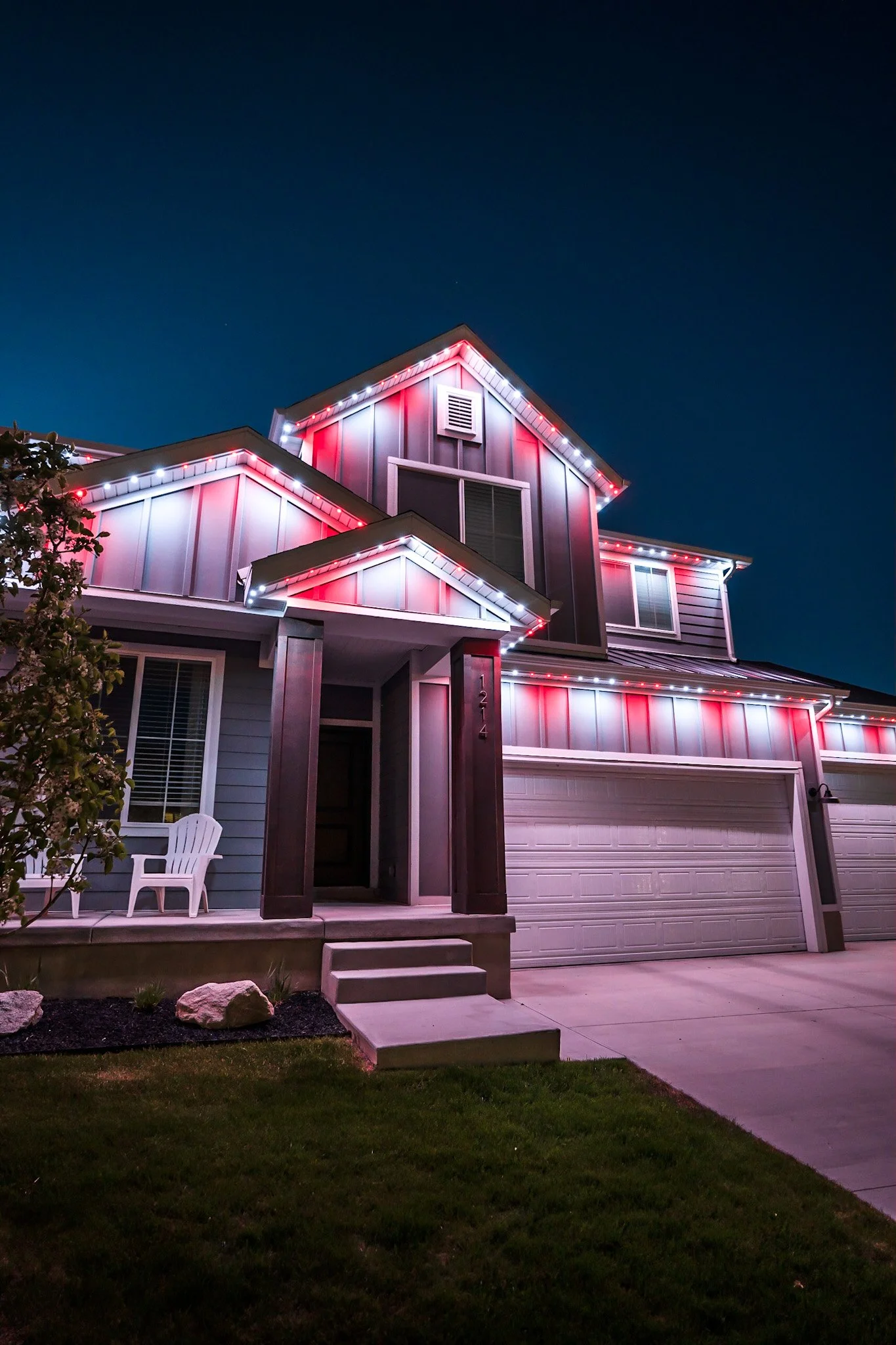 A modern two-story house at night decorated with red and white Christmas lights outlining the roof and edges.