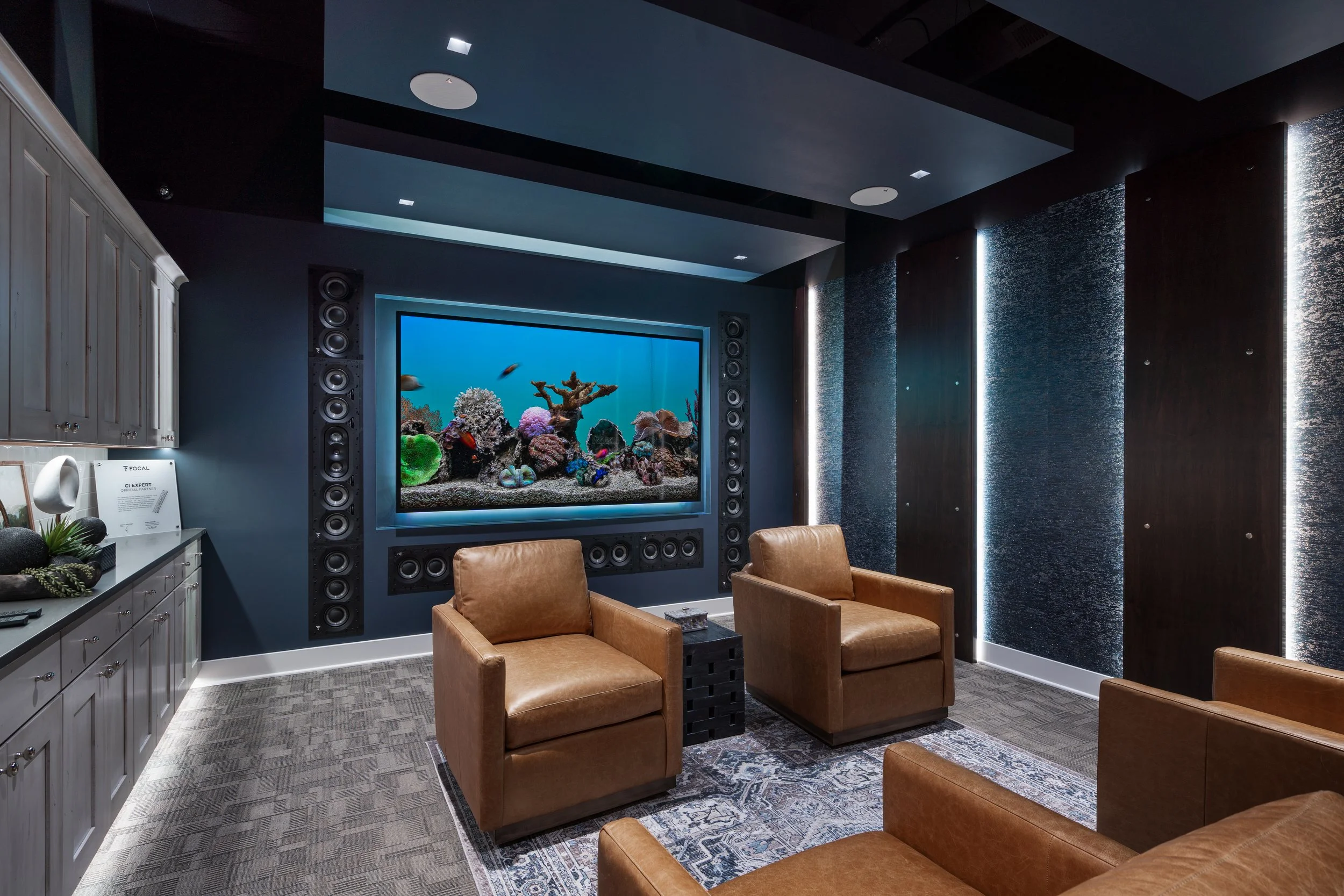 A room with dark walls and ceiling, brown leather armchairs, a large aquarium with colorful coral and fish, and speakers around the aquarium.