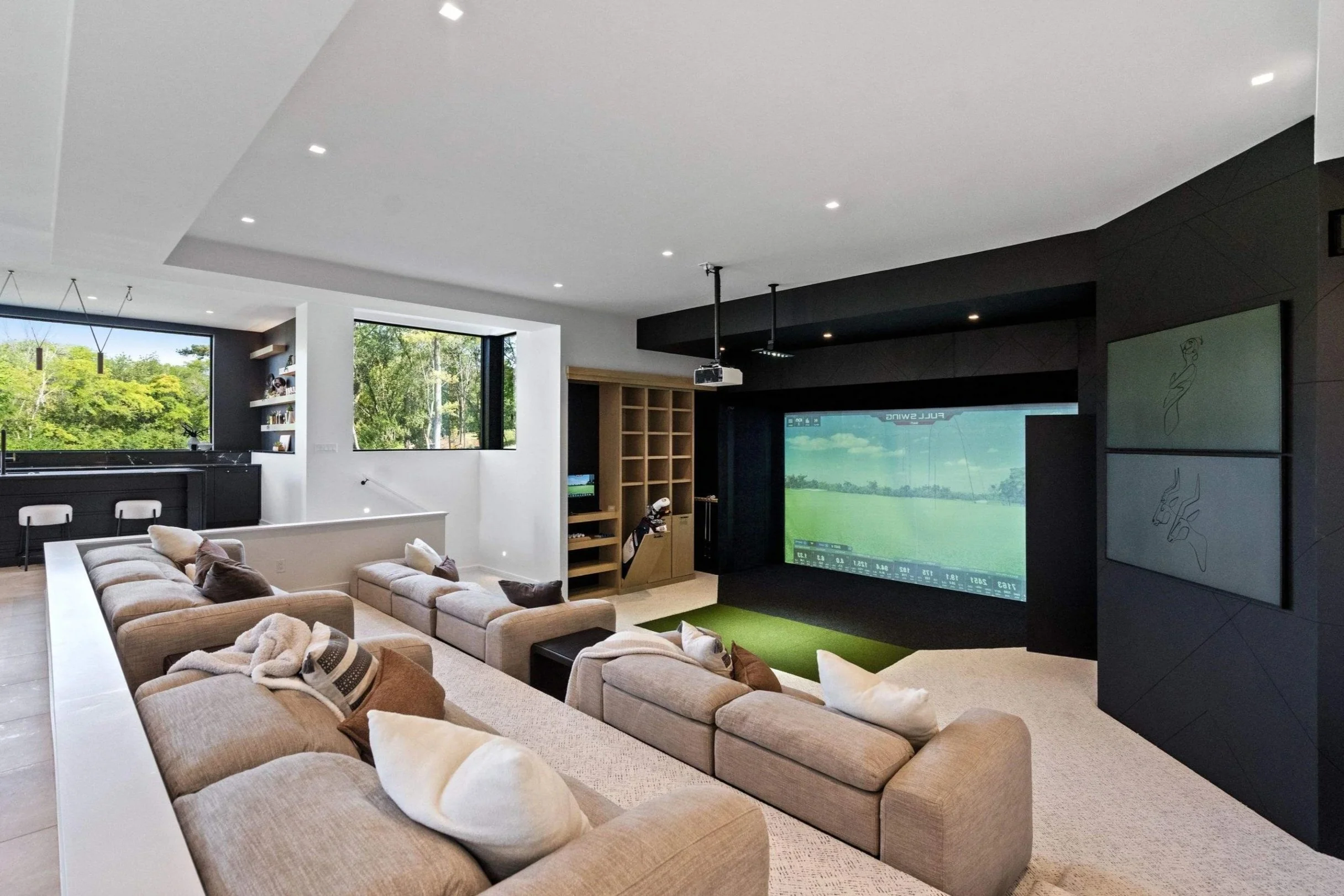Modern living room with sectional sofa, large screen golf simulator, and a kitchenette near windows showing green trees outside.