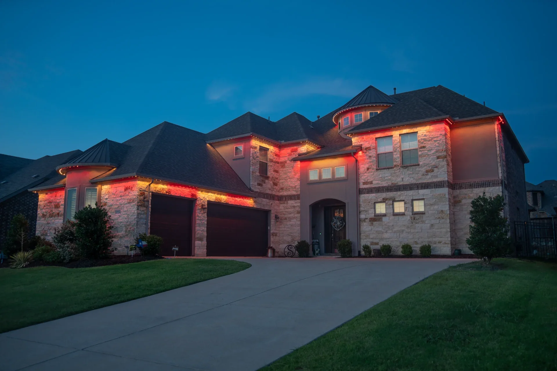 A large two-story house with stone and stucco exterior, illuminated by red and warm white lights, during dusk. The house features a dark shingled roof, multiple gables, and a driveway with a green lawn in front.