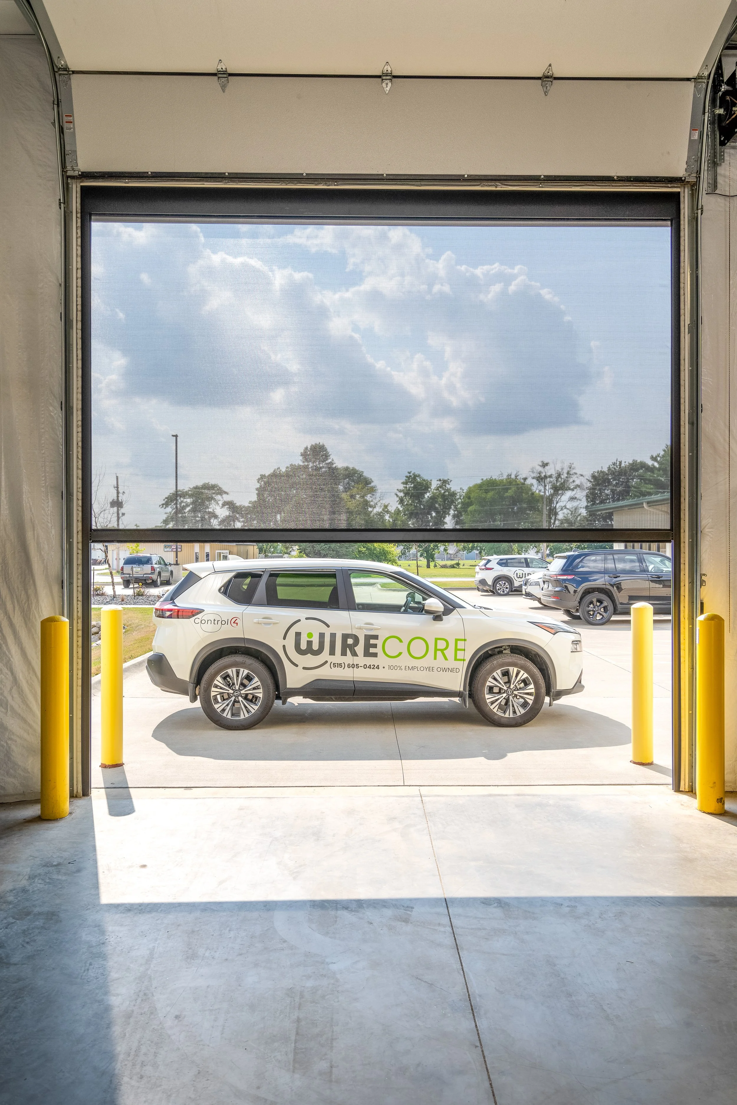 View of a white vehicle with WiredCore company logo parked outside a building, seen through a large open garage door with yellow safety bollards.