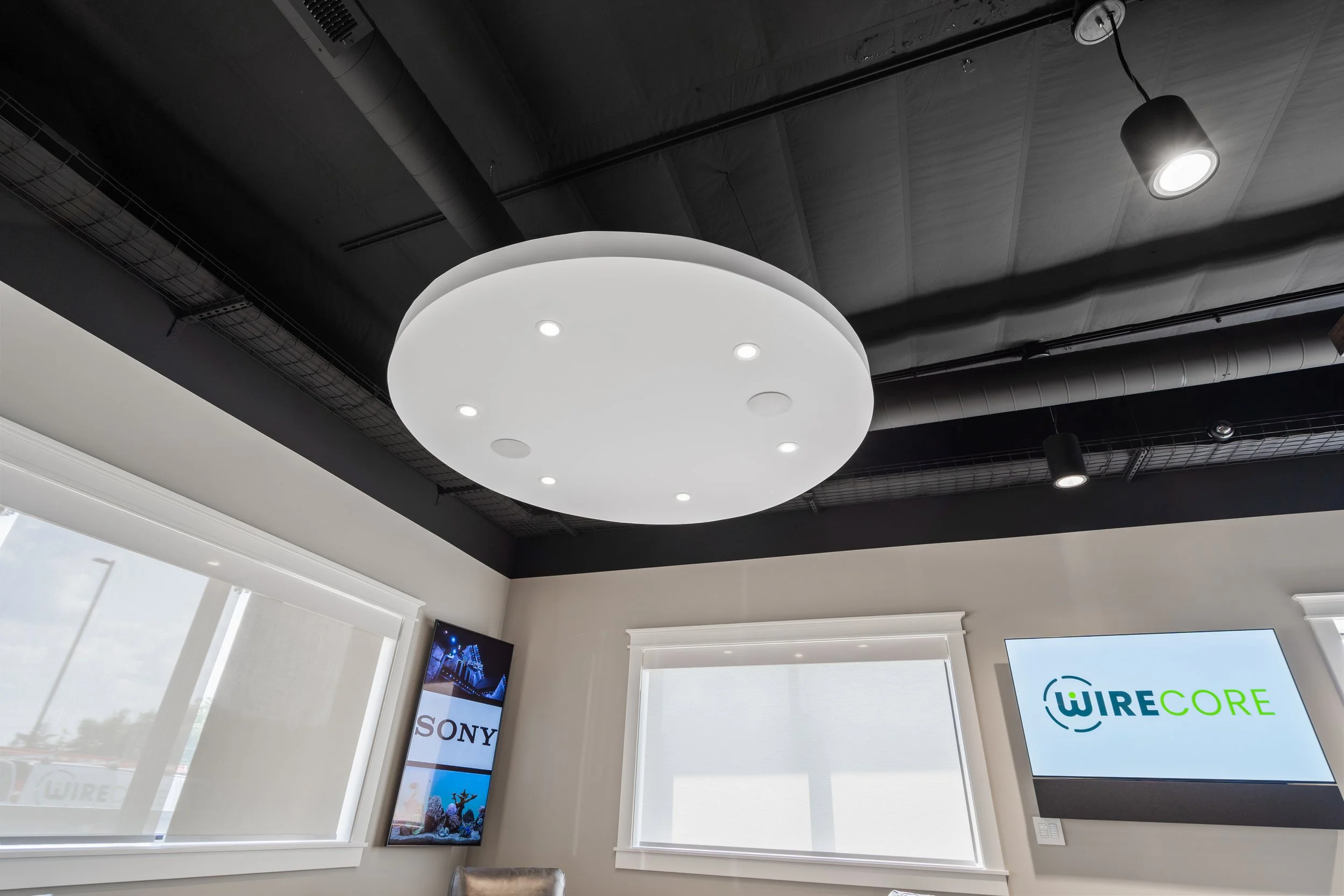 Interior office with a round white ceiling light, black ceiling panels, two windows with blinds, a wall-mounted TV displaying 'WIRE CORE' and 'SONY' logos, and modern track lighting.