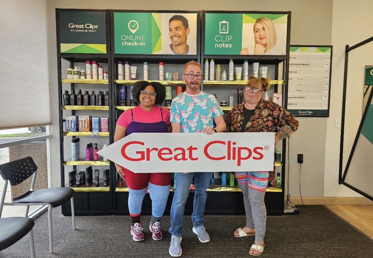 Three people standing inside a hair product store, holding a large arrow-shaped sign that says "Great Clips." They are smiling and standing in front of shelves filled with hair care products. There are signs above them advertising online check-in and