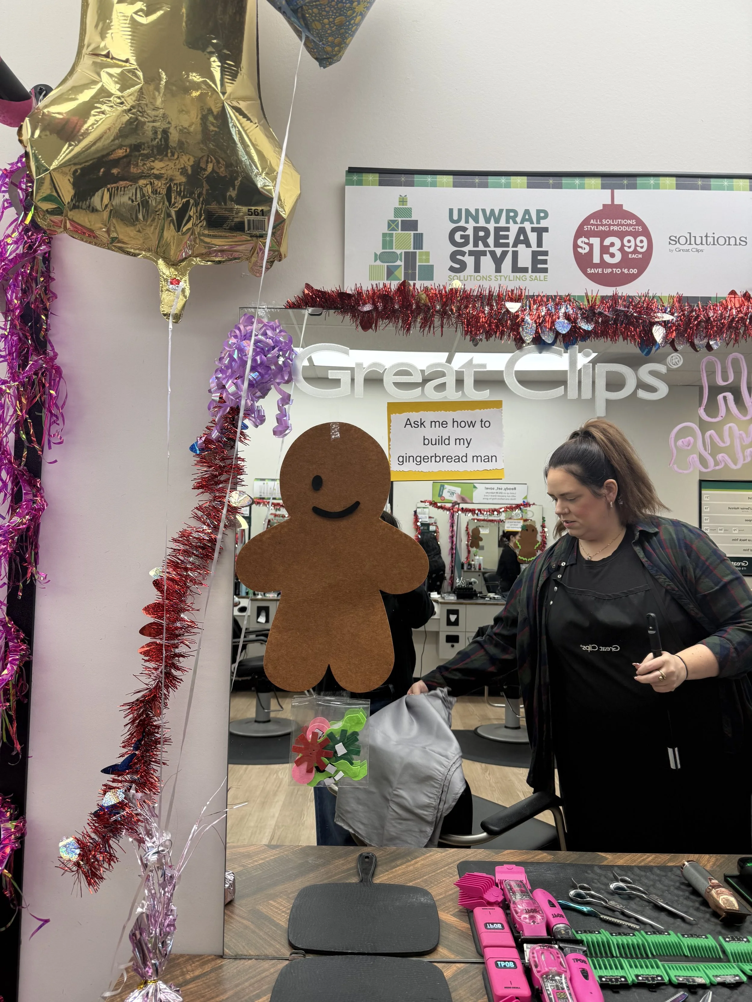 A salon station decorated with holiday decorations, including balloons, tinsel, and a gingerbread man cutout hanging in front of a mirror. There are hair styling tools on the counter, and a person with brown hair tied back is looking at something in 