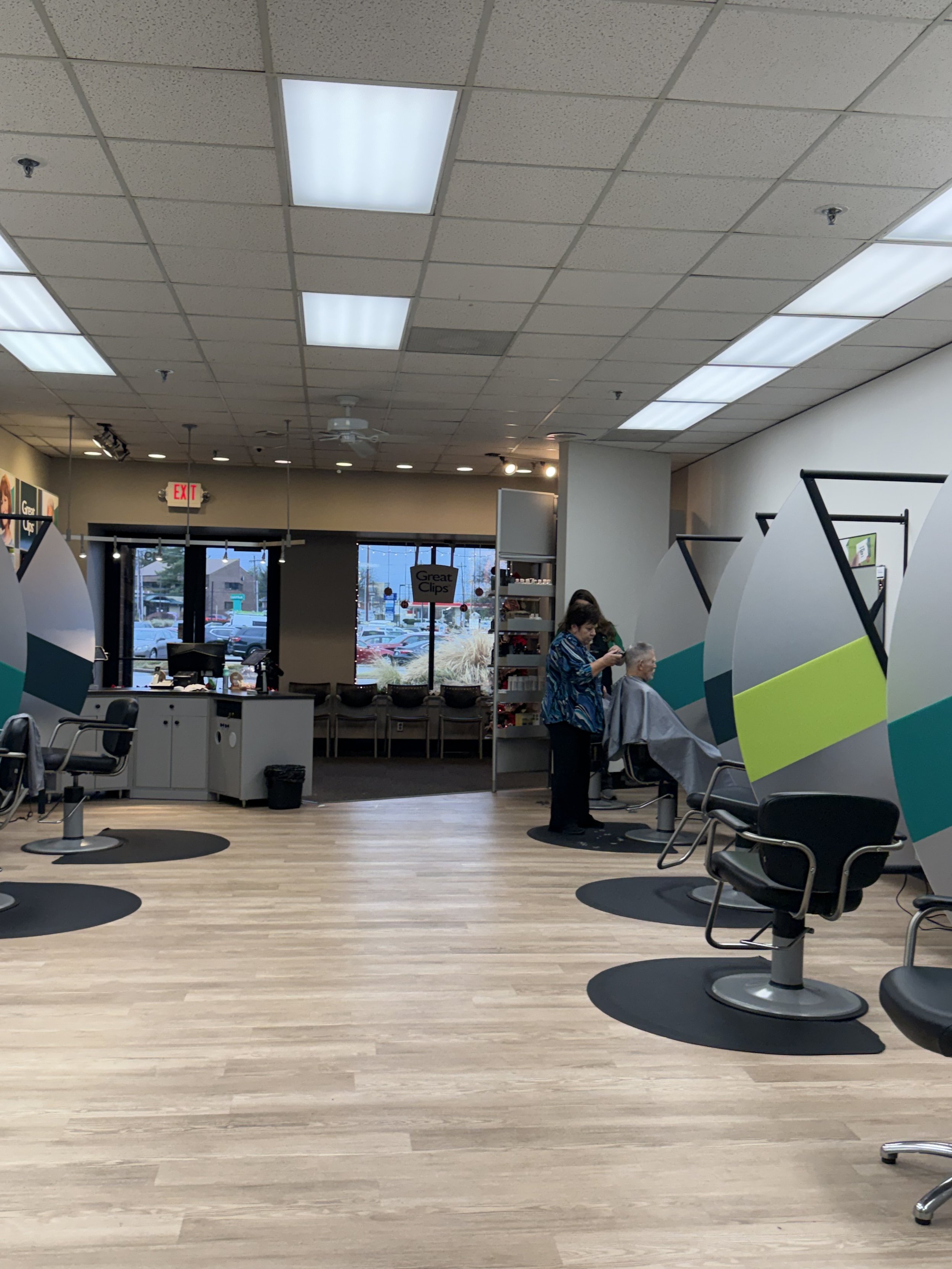 A woman getting her haircut in a salon with modern chairs, large mirrors, and a light wood floor. Two stylists are working on clients, and there are large windows and a seating area outside.