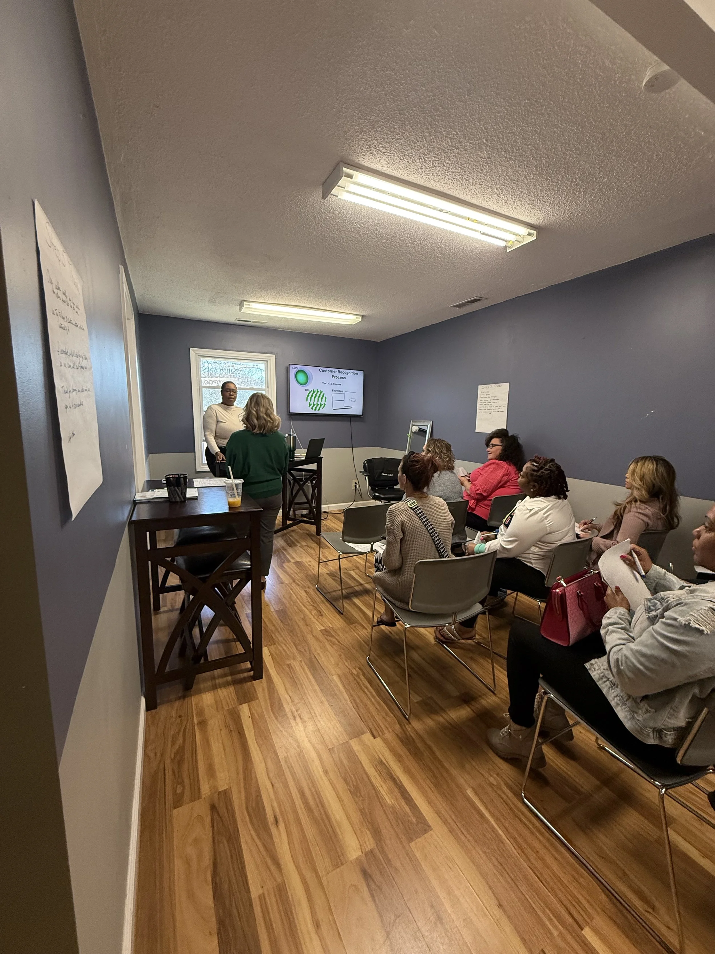 Group of women attending a presentation in a small conference room with blue walls, chairs, a laptop, and a large screen displaying a slide about customer recognition process.