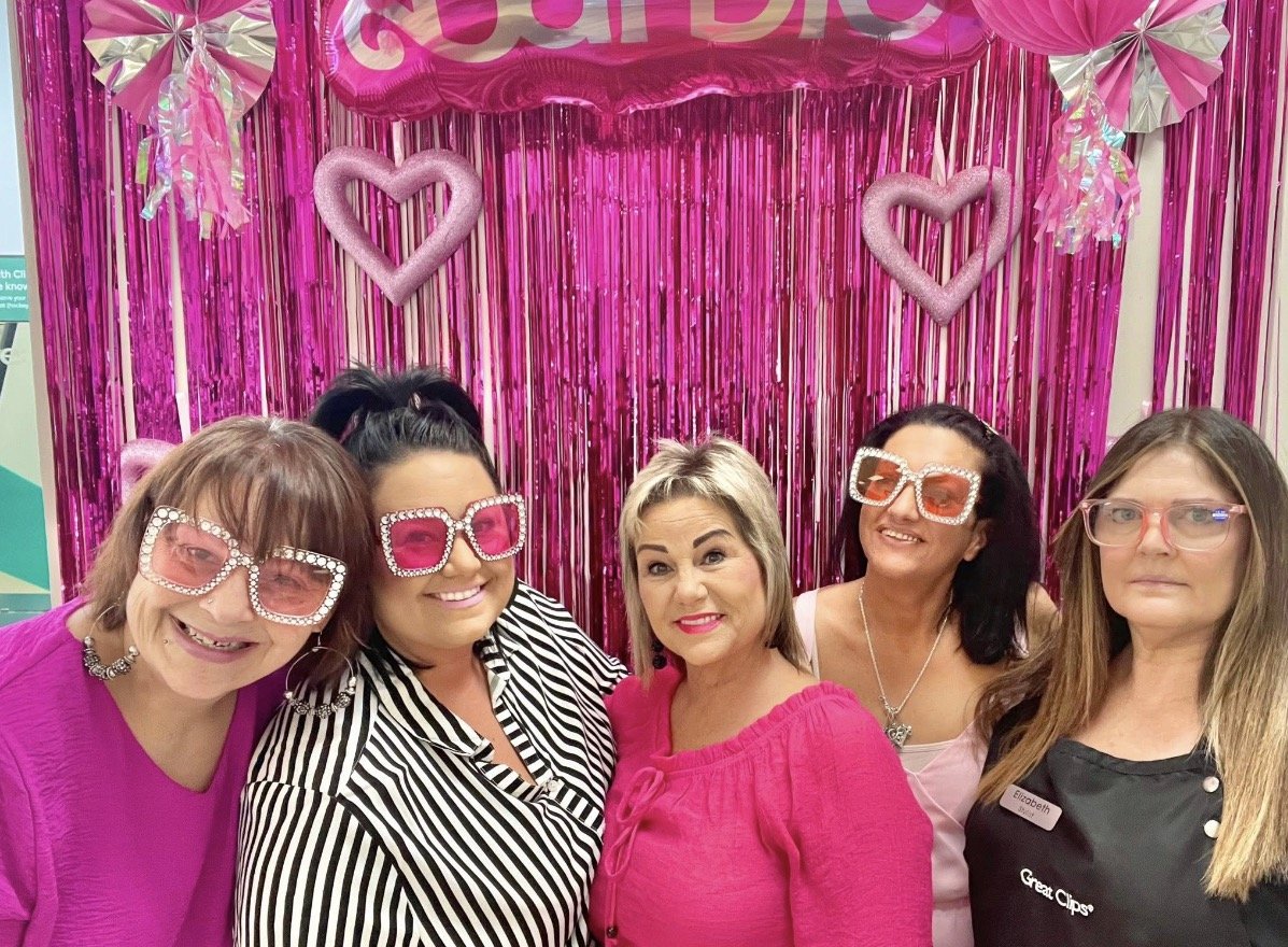 Group of five women wearing pink and black outfits with oversized pink and rhinestone sunglasses, standing in front of pink decorations with hearts and metallic streamers.