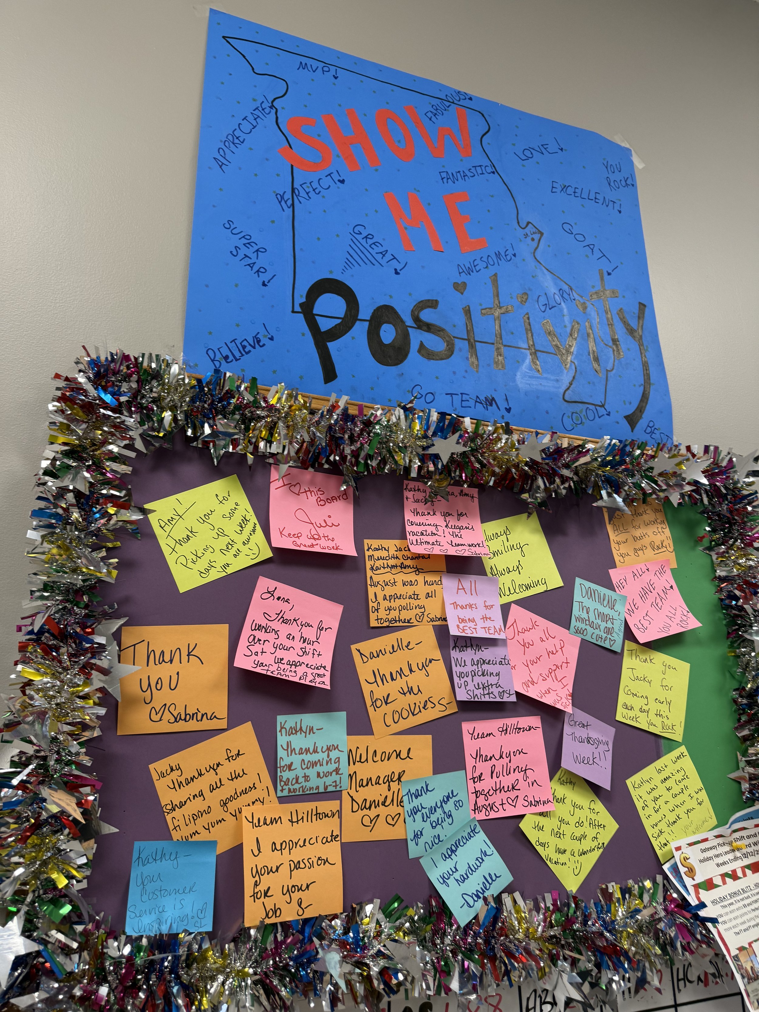 Decorated bulletin board with many colorful sticky notes of thanks and appreciation, surrounded by festive tinsel. A large blue poster at the top reads 'Show Me Positivity' in bold letters.