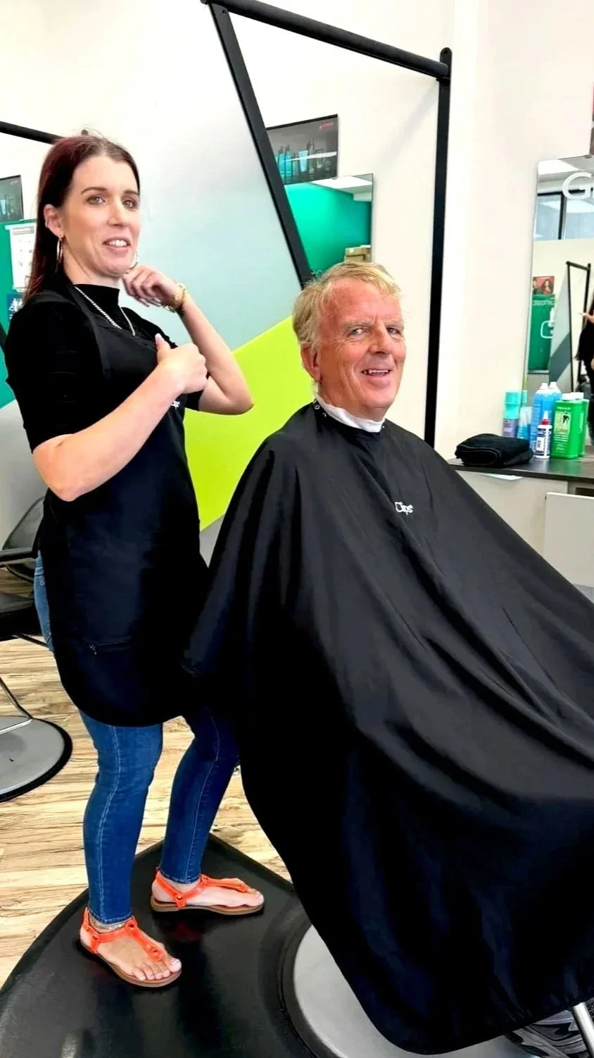 A woman at a hair salon giving a man a haircut while standing on a balance board.