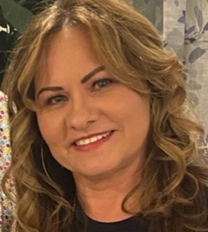 A woman with light brown hair and earrings taking a selfie inside a car, with a necklace and a black top.