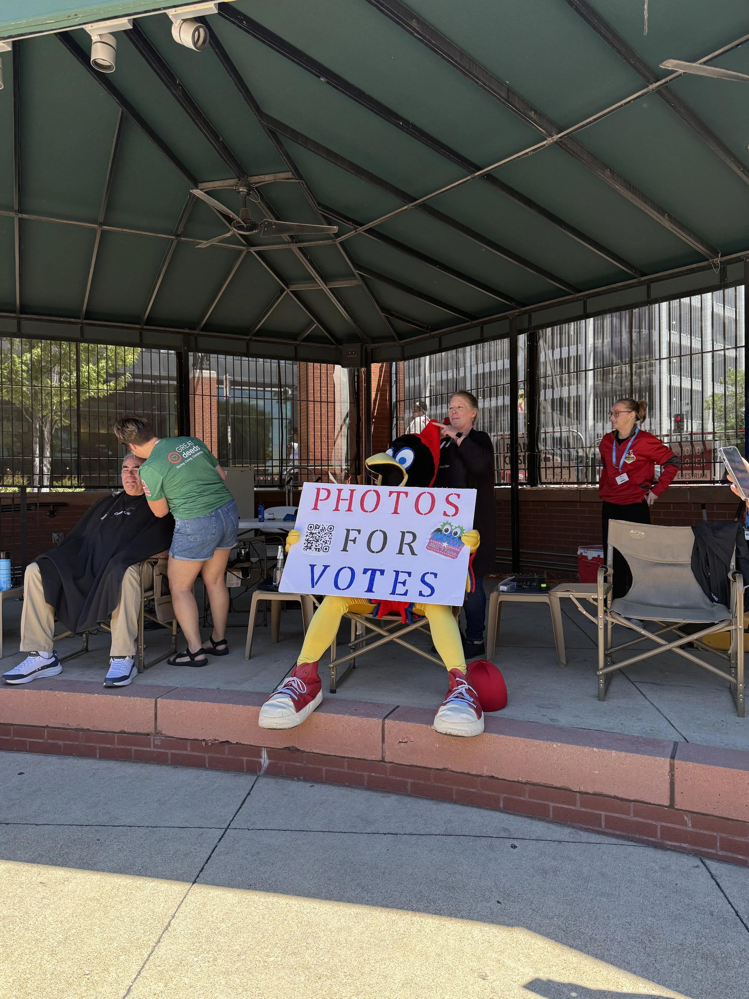 A person getting a haircut under a covered outdoor pavilion, with a person in a chicken mascot costume holding a sign that says 'Photos for Votes', and two women standing nearby, one with a phone.