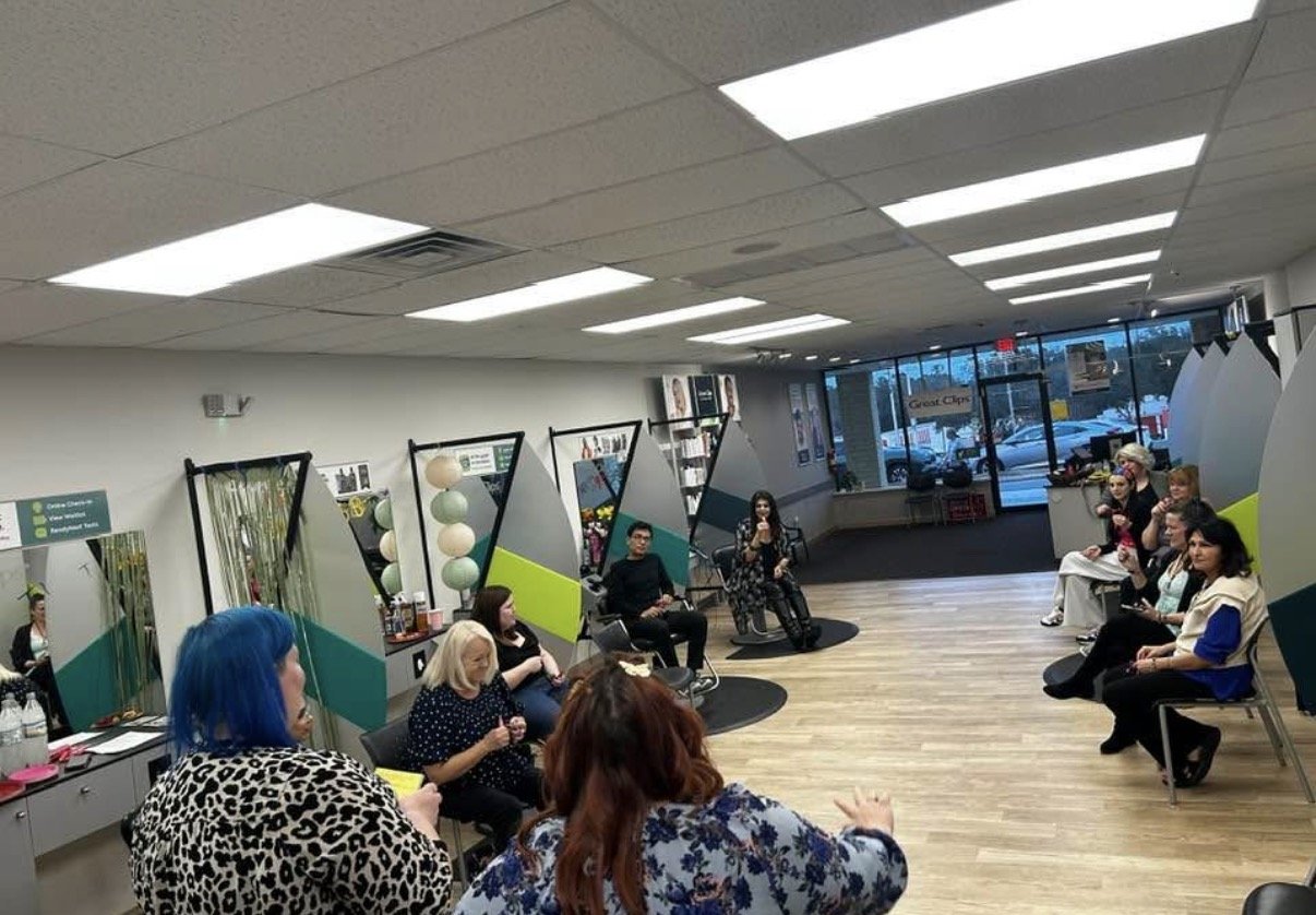 A group of people seated in a hair salon, participating in a class or seminar, with styling stations and large windows in the background.