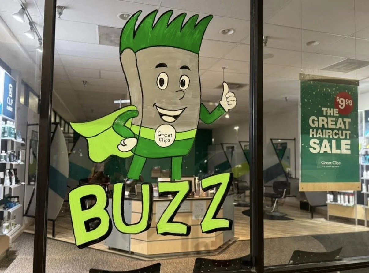 Cartoon character of a buzz cut hairstyle with green hair, wearing green superhero costume with a cape, giving a thumbs-up sign, with words "BUZZ" and a badge saying "Great Clips" on it, on a glass window of a hair salon, with a poster advertising a 