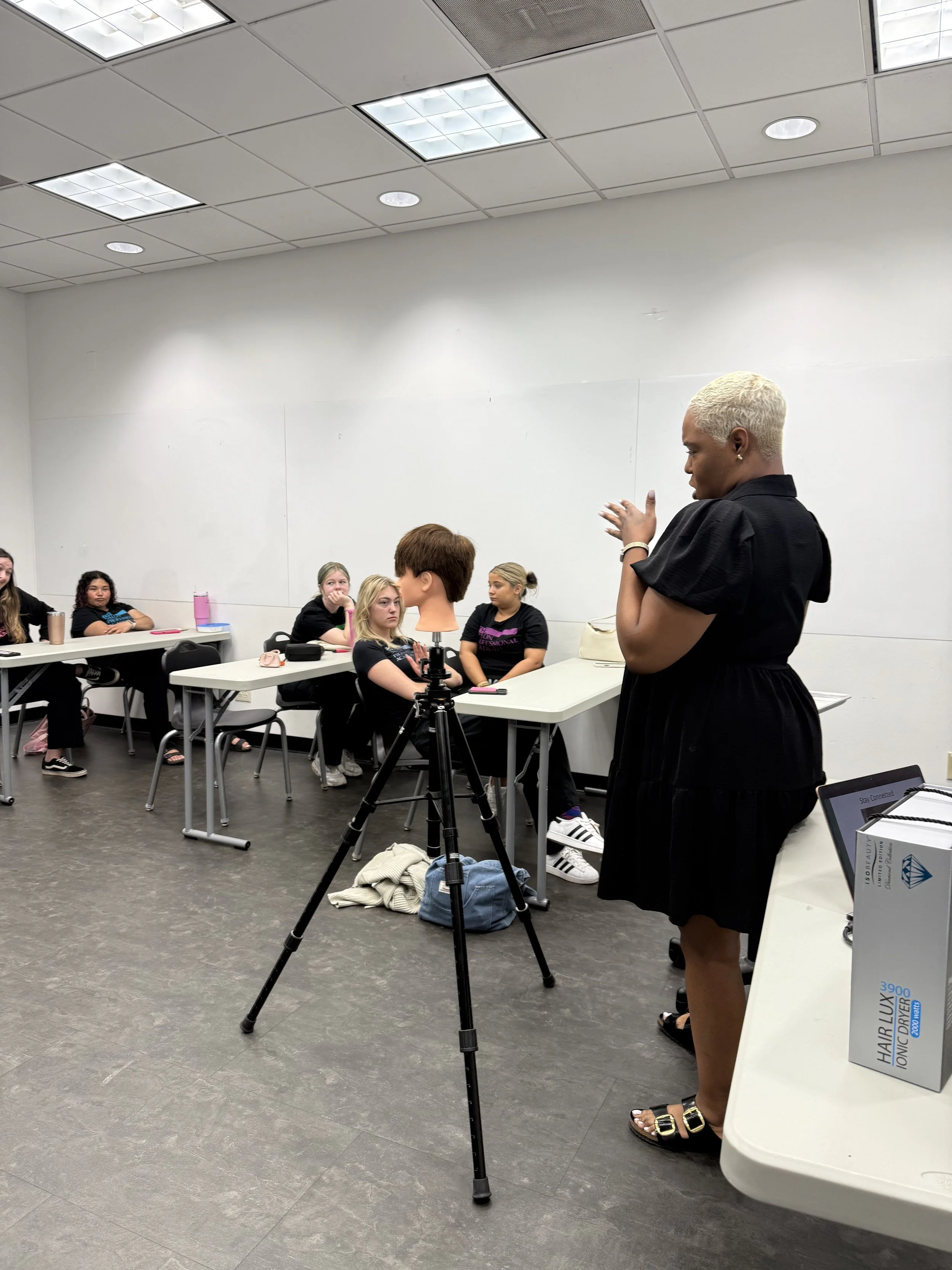 A classroom or workshop setting with a woman demonstrating hair styling techniques to a group of students. The students are seated at tables and watching her attentively.
