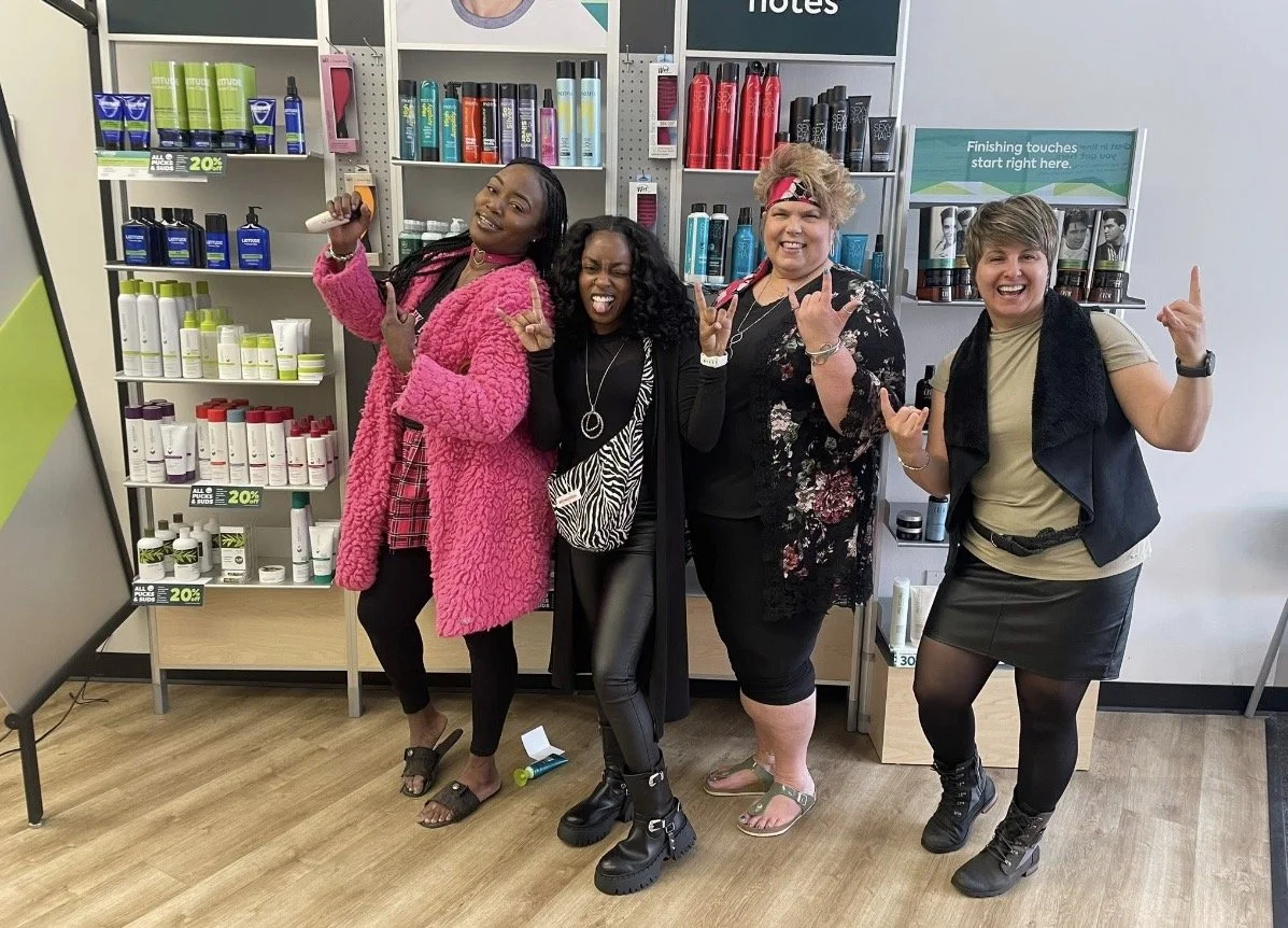 Four women posing cheerfully in a store aisle with hair and beauty products on shelves behind them. They are making rock and peace signs and smiling.
