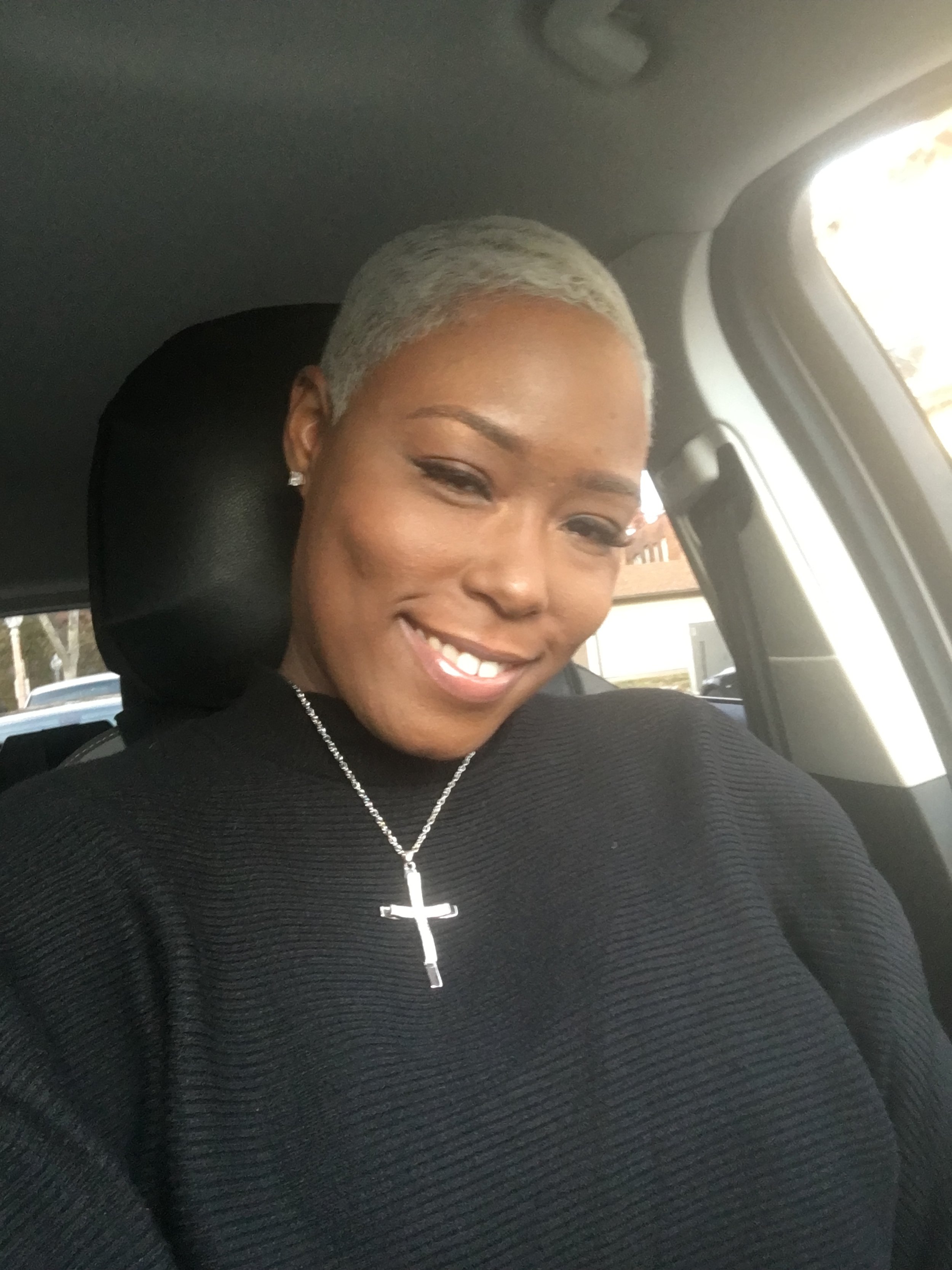 A smiling woman with short, platinum blonde hair sitting in a car, wearing a black top, a silver cross necklace, and stud earrings.