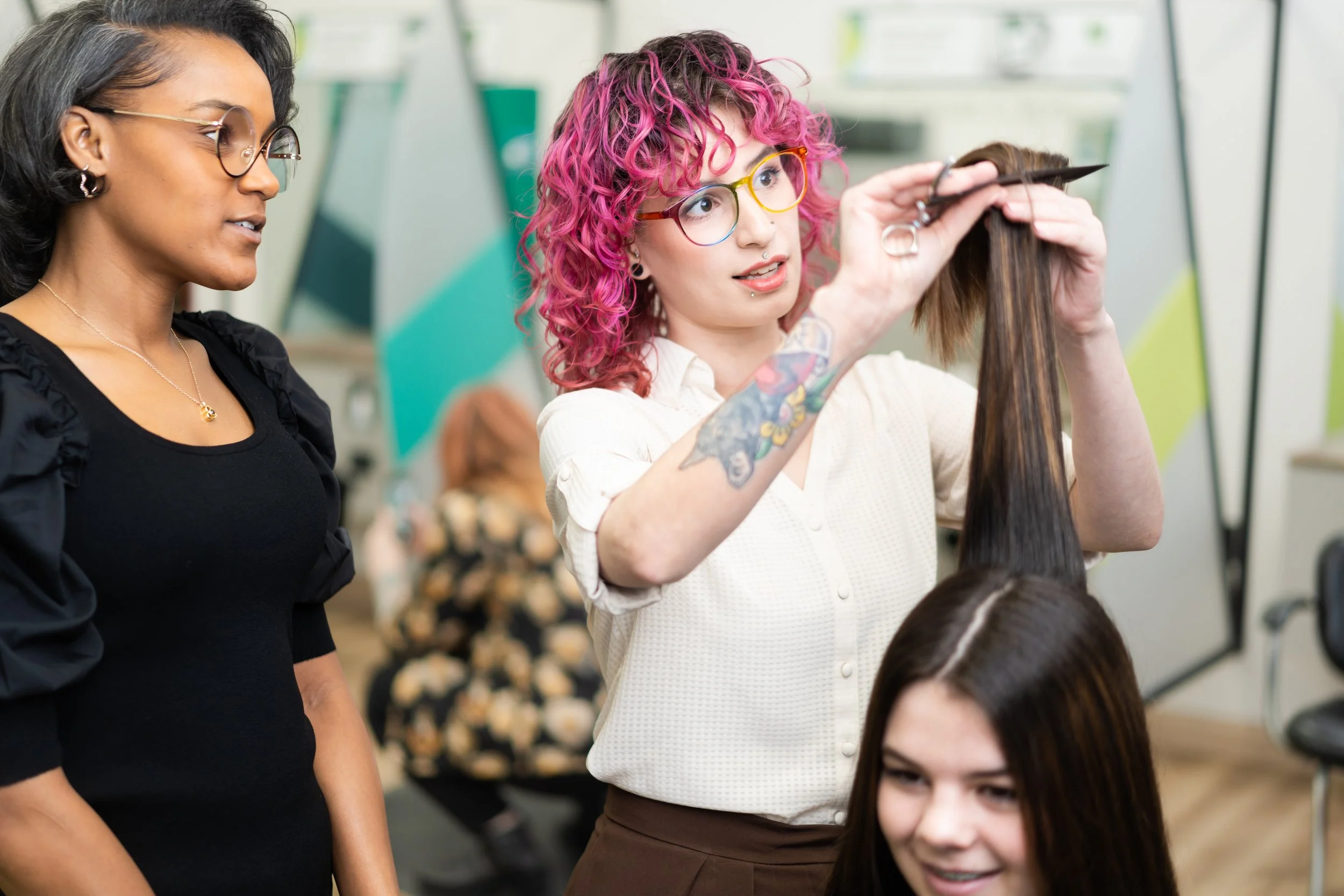Hairdresser with pink curly hair and glasses demonstrating haircut techniques to a client in a salon.