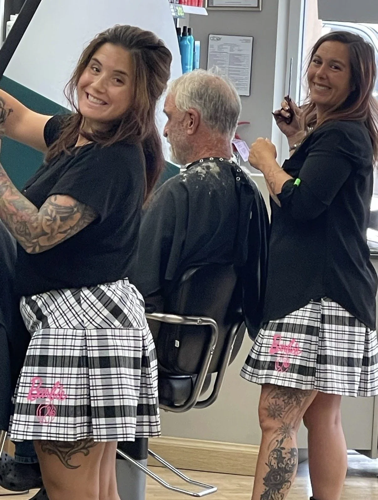 Two women with tattoos wearing black t-shirts and plaid skirts are standing next to a man getting a haircut at a barbershop. The women are smiling and holding scissors, while the man is seated with a barber cape on.