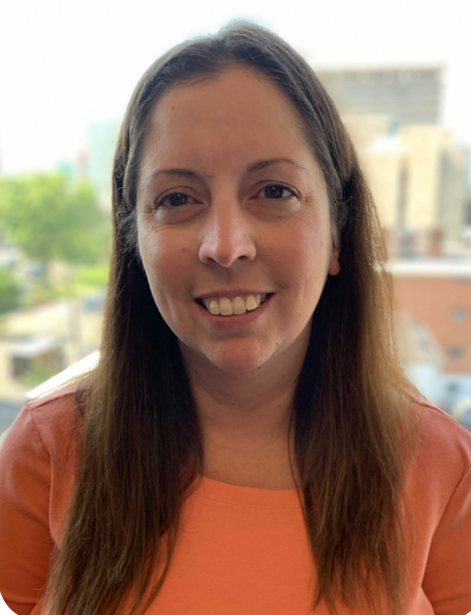 A woman with long brown hair smiling, wearing a coral-colored shirt, with a cityscape and green trees in the background visible through a window.