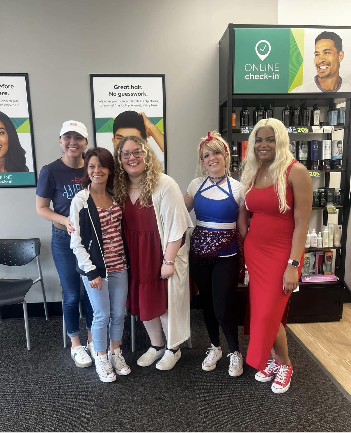 Five women standing together inside a retail store, smiling for a group photo. The store has signs promoting online check-in and product displays.