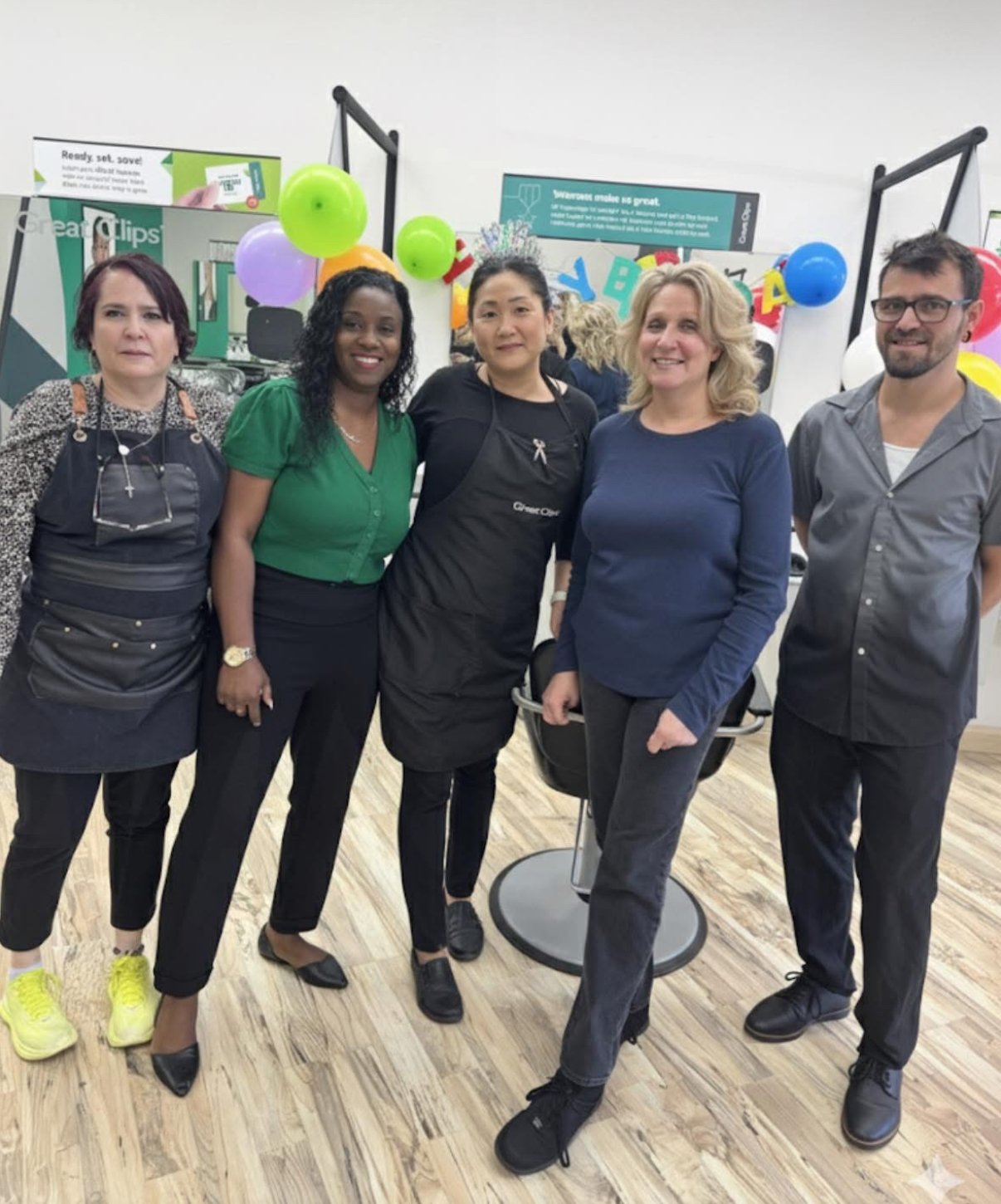 Five people standing together celebrating a birthday, with colorful balloons and party decorations in the background.