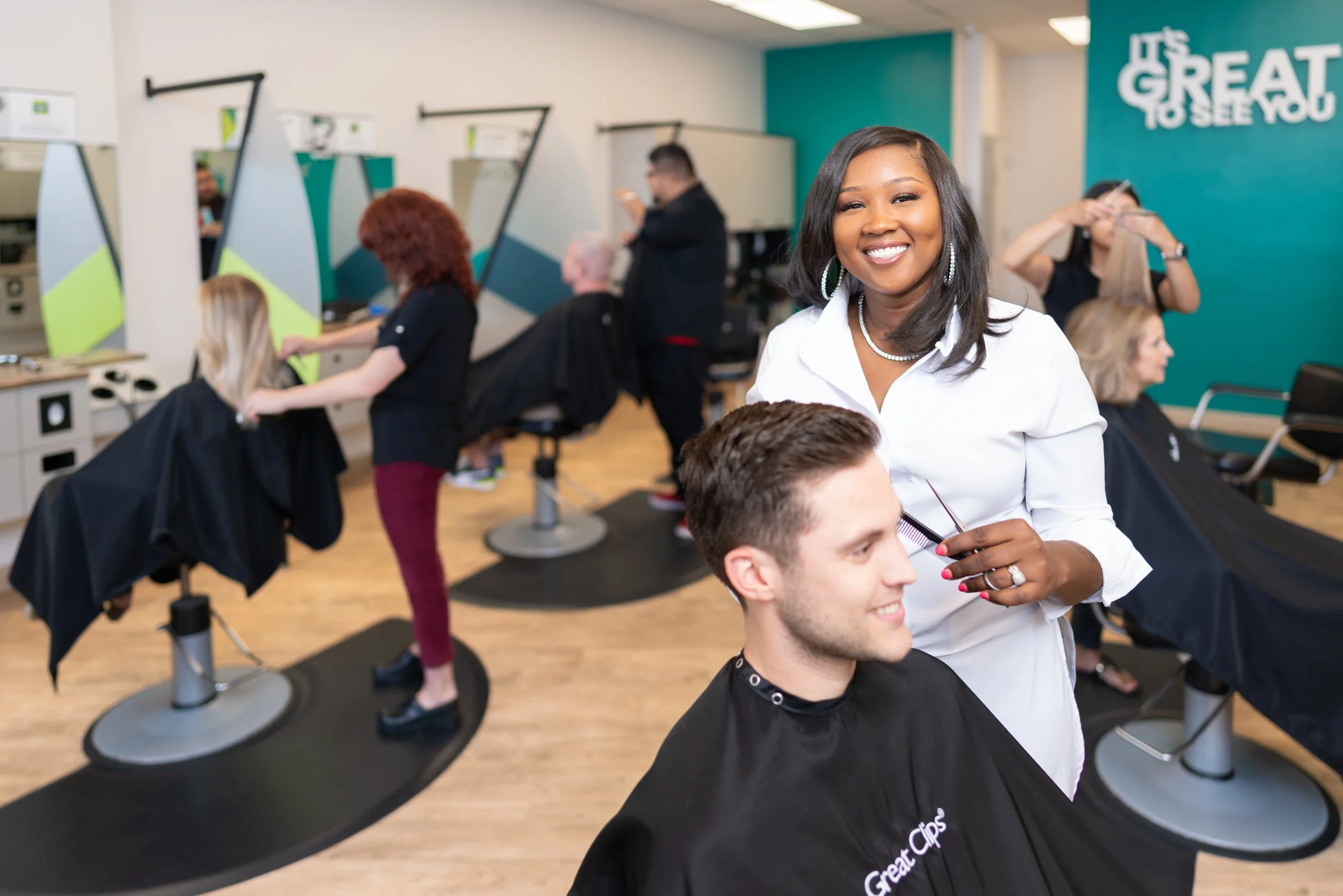 A smiling woman dressed in white and holding glasses, standing beside a man with short dark hair, who is seated with a barber's cape at a hair salon. Several other clients and hairstylists are visible in the background, working on haircuts.