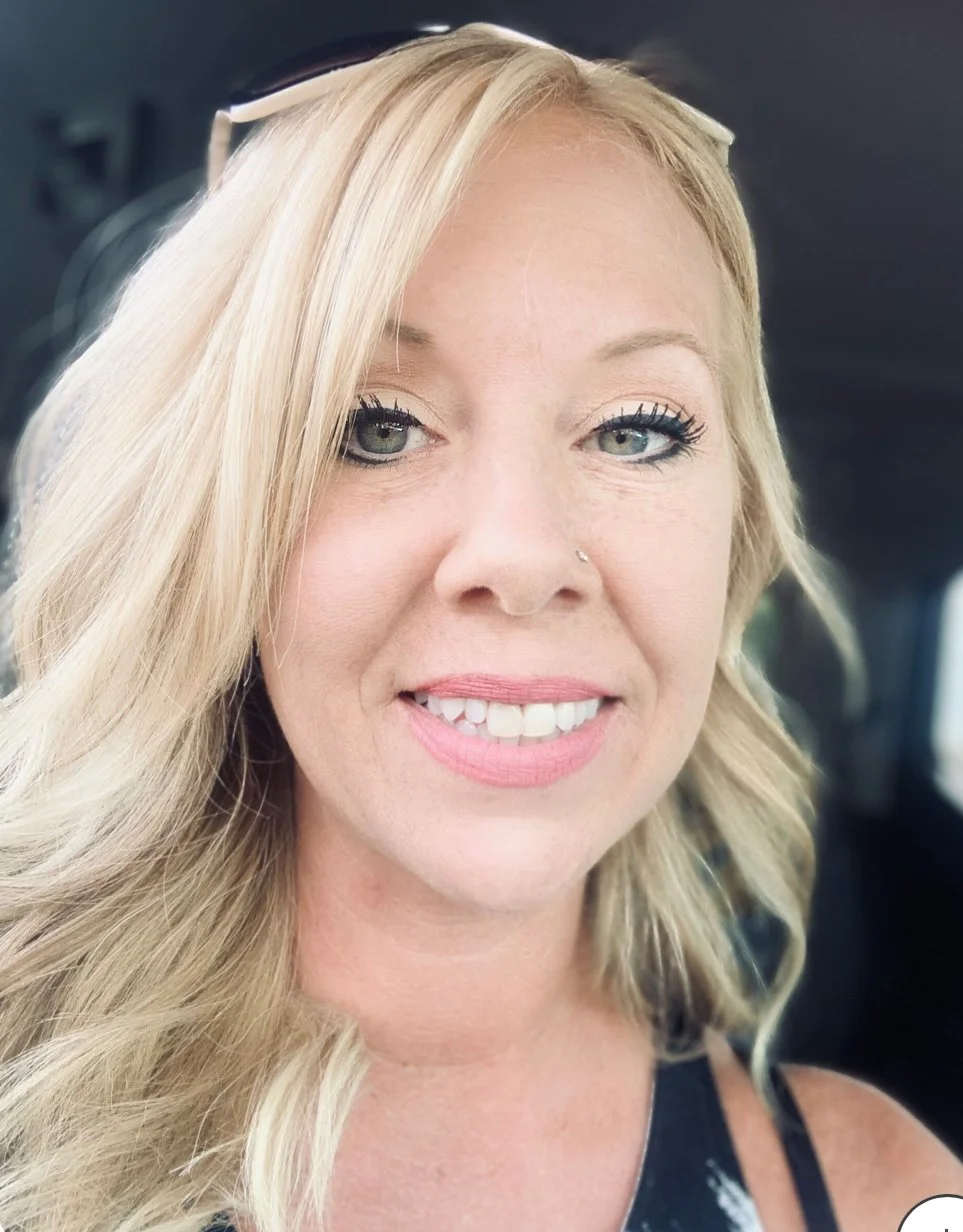 Close-up of a woman with blonde hair, light makeup, and a nose piercing, smiling in a car.