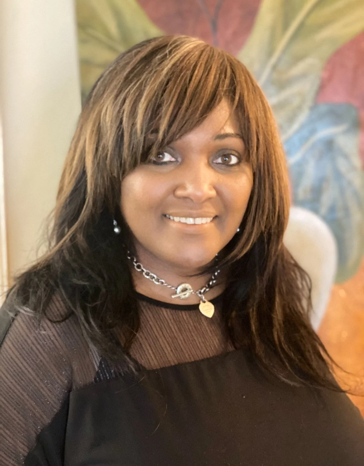 A woman with shoulder-length brown hair, wearing a black top with sheer sleeves, and jewelry including earrings, a layered necklace, and a heart-shaped pendant, smiling in front of a colorful abstract background.