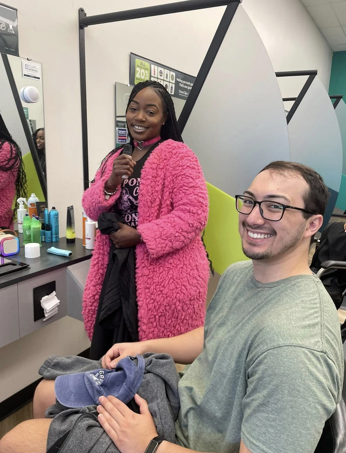 A woman with braids and a pink fuzzy coat standing next to a smiling man with glasses, holding a hat and clothes, standing in front of a mirror and a vanity with various toiletry bottles.