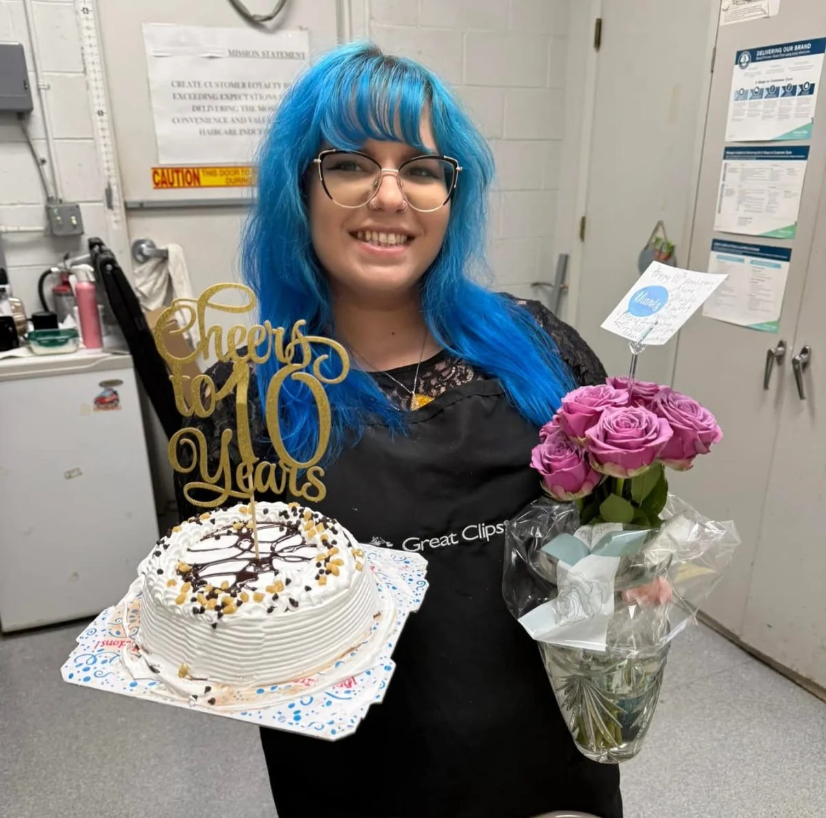 A woman with bright blue hair and glasses holding a birthday cake, a bouquet of pink roses, and a card, celebrating a 10-year milestone in a kitchen or break room.