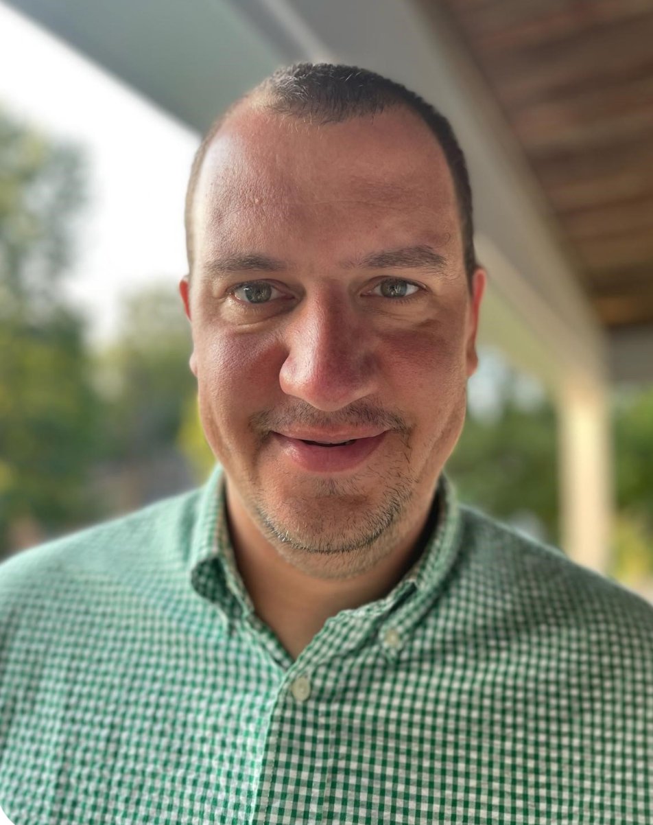 Close-up of a smiling man with short dark hair, light skin, wearing a green checkered shirt, standing outdoors with a blurred background of trees and a wooden porch roof.