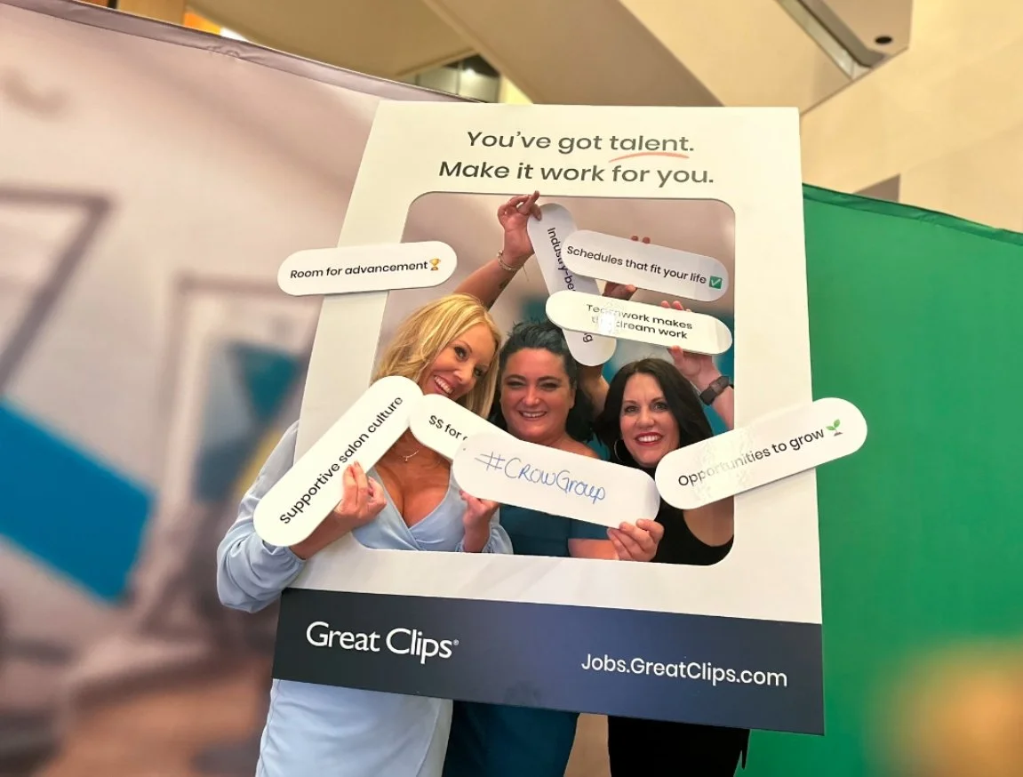 Three women smiling and holding a large photo frame prop with various messages and hashtags, promoting a positive work environment and personal growth.
