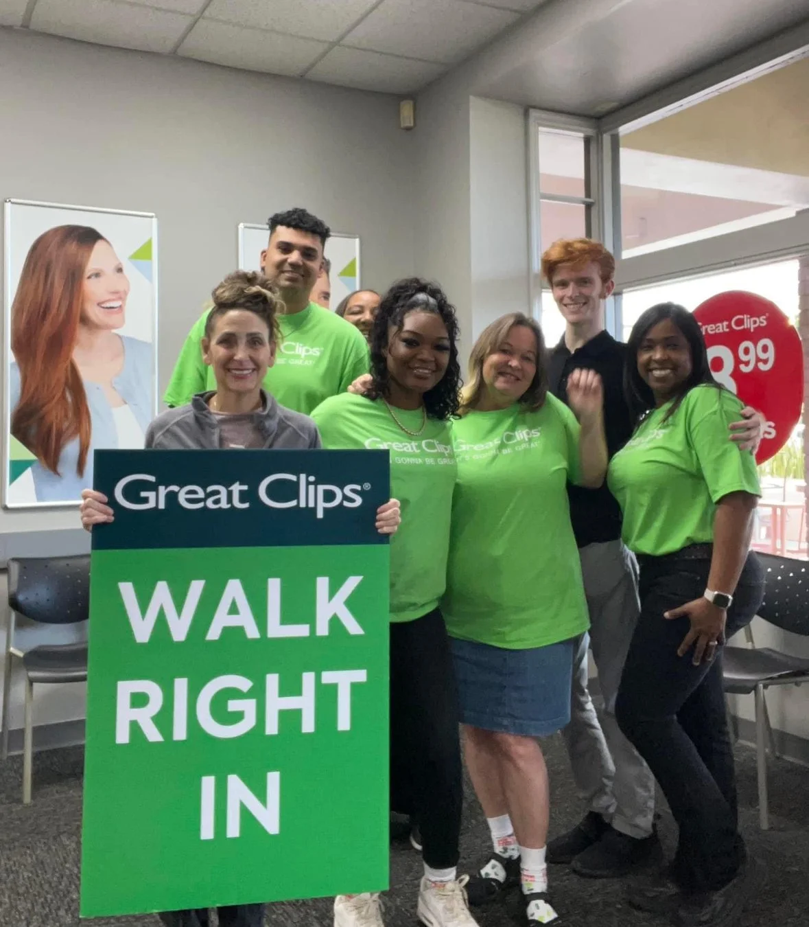 Group of people wearing green Great Clips T-shirts standing inside a salon near a large window. One person is holding a green sign that says 'Great Clips WALK RIGHT IN'. They are smiling and posing for the photo.