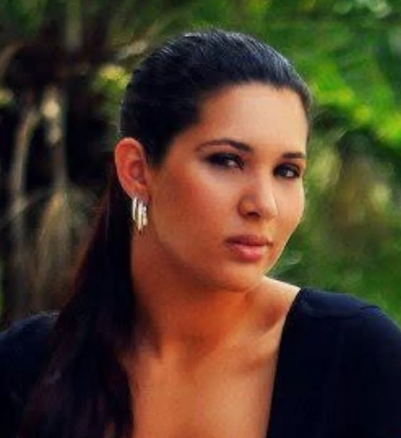 A woman with dark hair pulled back, wearing earrings, looking at the camera, outdoors with greenery in the background.