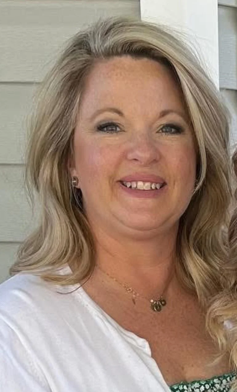 A woman with blonde, wavy hair smiling at the camera, wearing a white top and jewelry, standing in front of a light-colored wall.