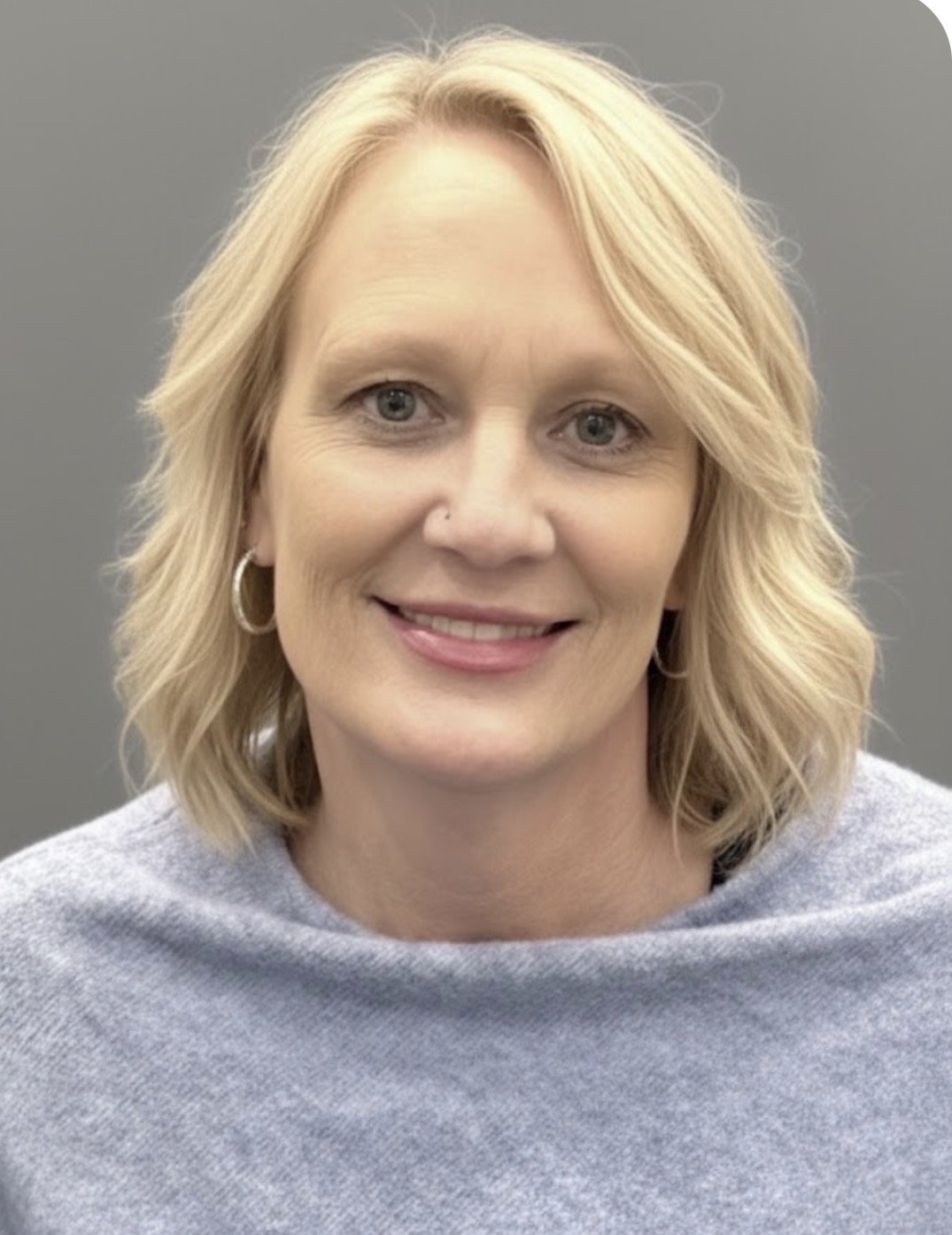 A smiling woman with blonde hair taking a selfie at a dining event, with a man partially visible behind her, and a table with food and drinks in the background.