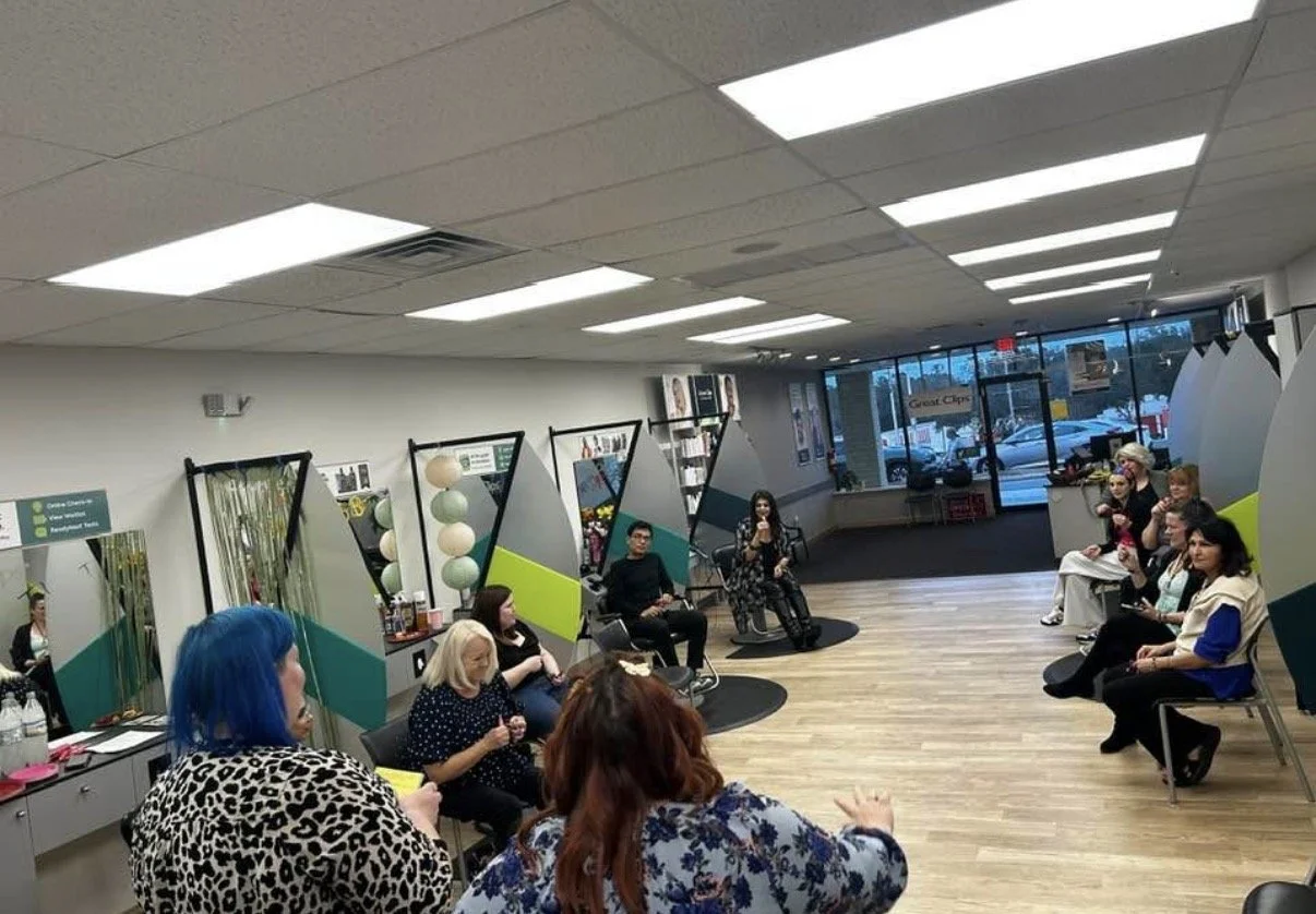 A group of people seated in a waiting area of a hair salon with styling chairs and mirrors, some using their phones or talking.