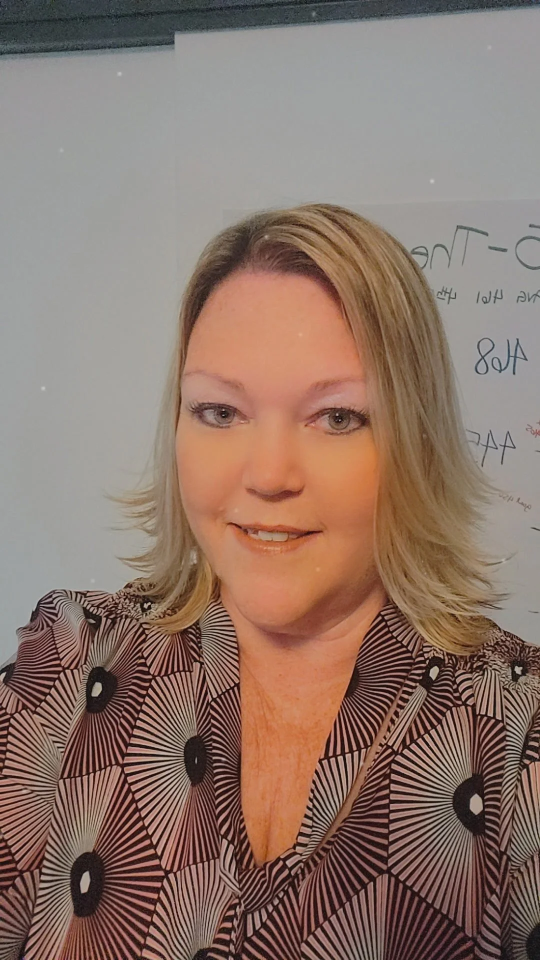 A woman with blonde hair and light-colored eyes smiling in front of a whiteboard with handwritten notes.