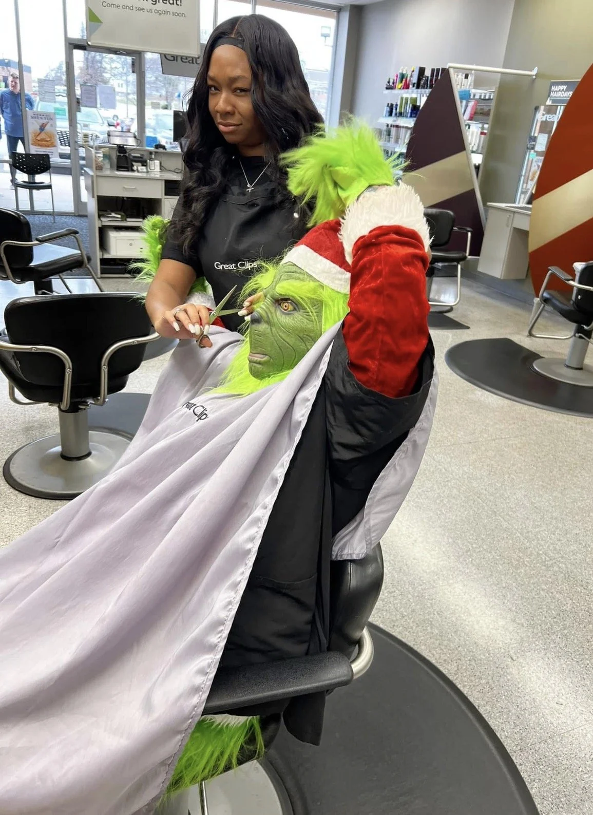 A person with a Vegita-like face and green hair, dressed in a Santa hat, has their hair cut at a hair salon. A hairstylist stands beside them holding scissors, preparing to cut their hair.