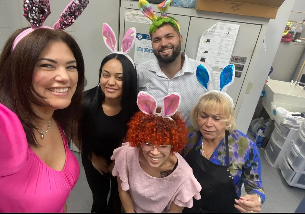 Group of five people celebrating Easter, wearing bunny ears hats, in an office or kitchen setting. They are smiling and posing for a selfie.