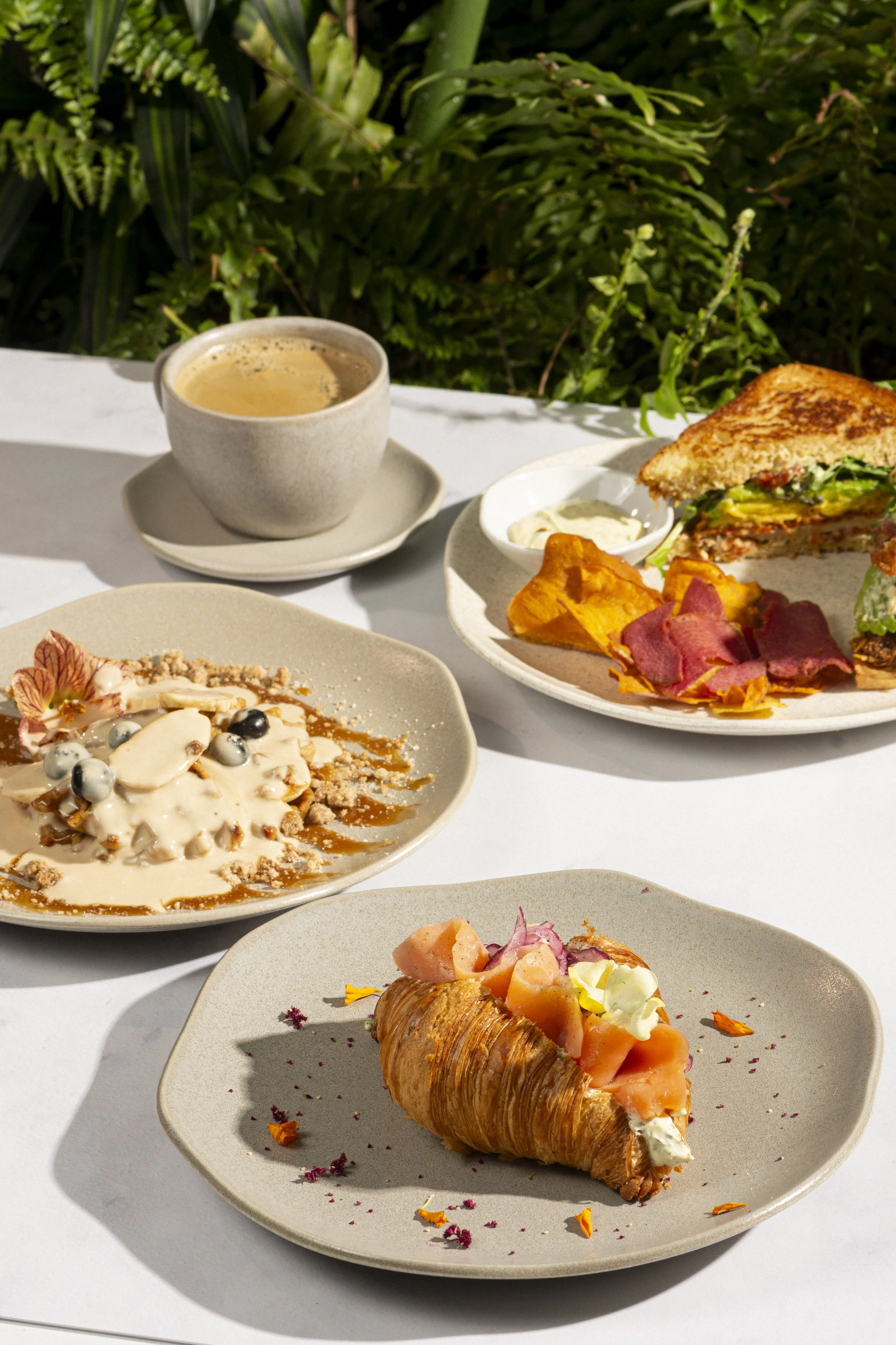 Mesa de desayuno con café, sándwich, postre de crema y fruta, y un croissant con frutas y crema, en un ambiente natural.