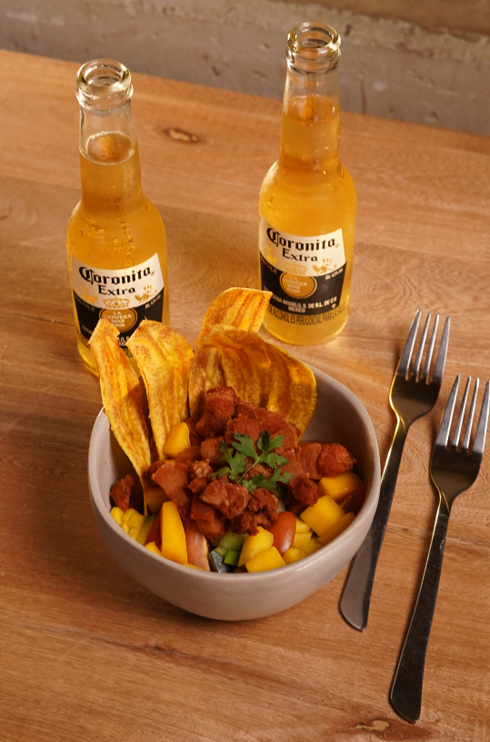 Plato con carne en trozos con fruta y tostones, acompañado de dos cervezas Corona y cubiertos en una mesa de madera.