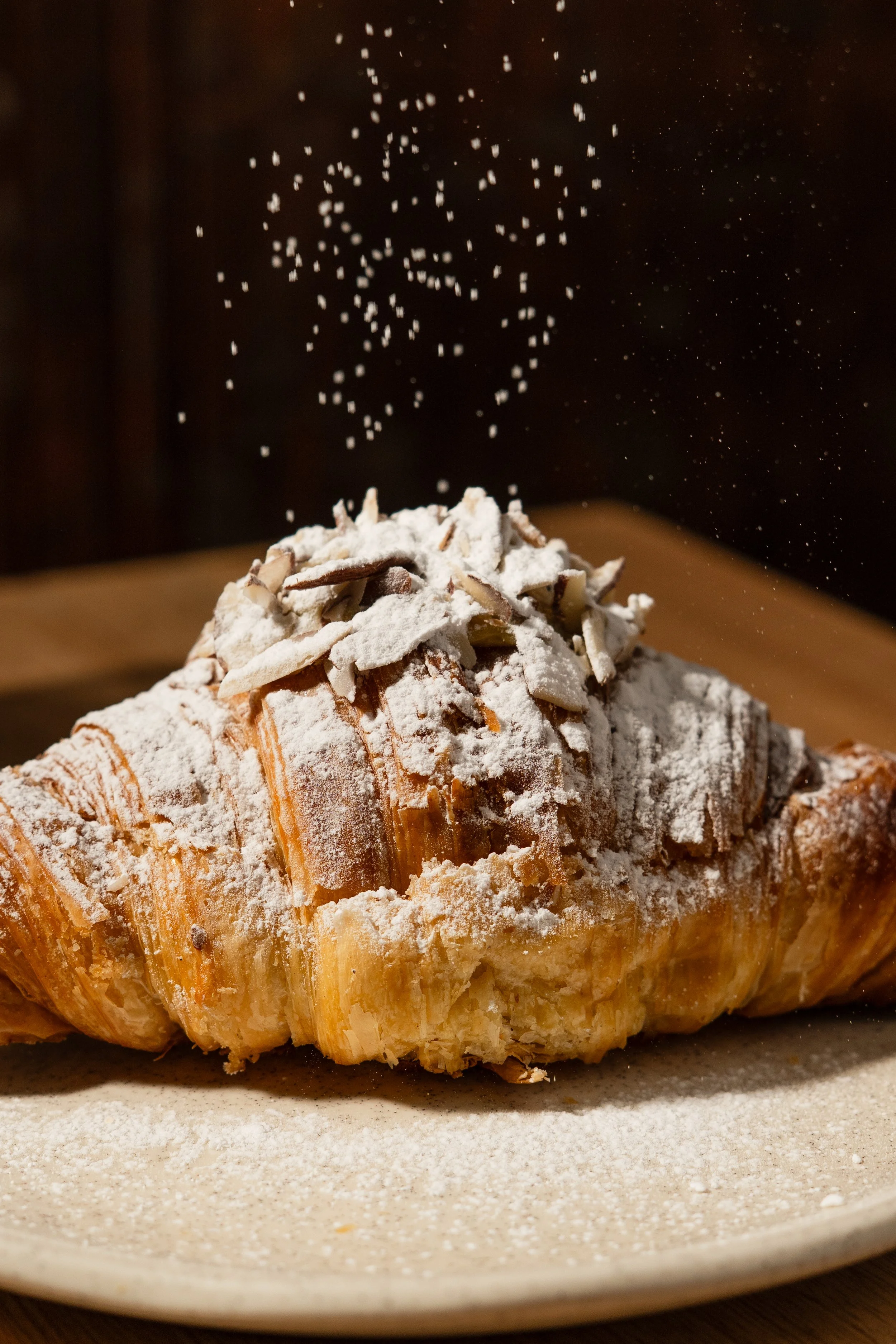 Un croissant cubierto de azúcar en polvo y almendras en una placa, con azúcar en polvo cayendo sobre él.