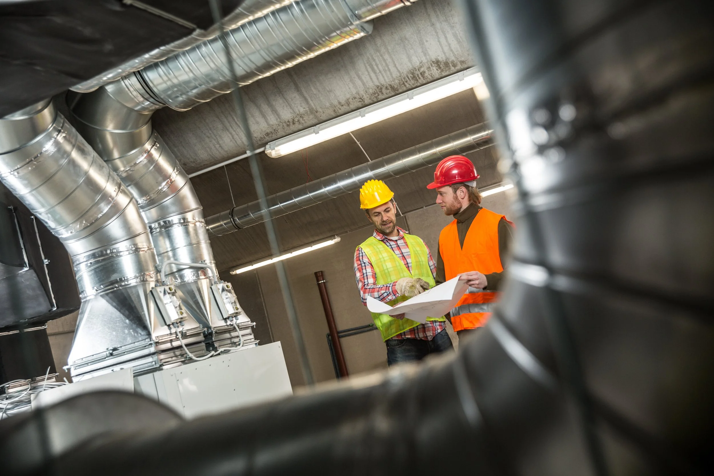 Two workers wearing safety helmets and vests reviewing a blueprint in an industrial setting with metal ducts and pipes.