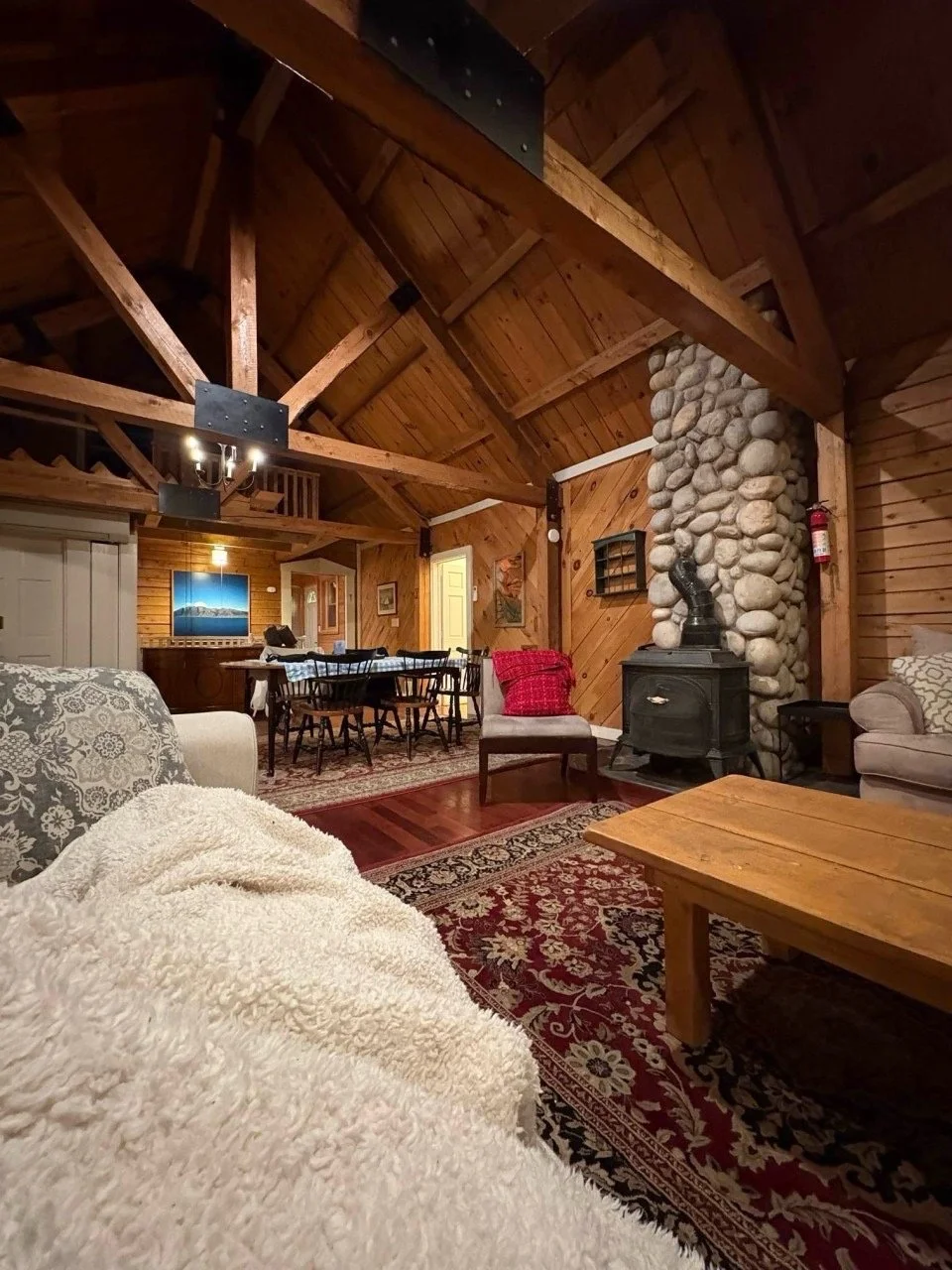 Interior of a cozy cabin living room with wooden walls and ceiling, a stone fireplace, couches, a wooden coffee table, a dining table with chairs, and warm lighting.