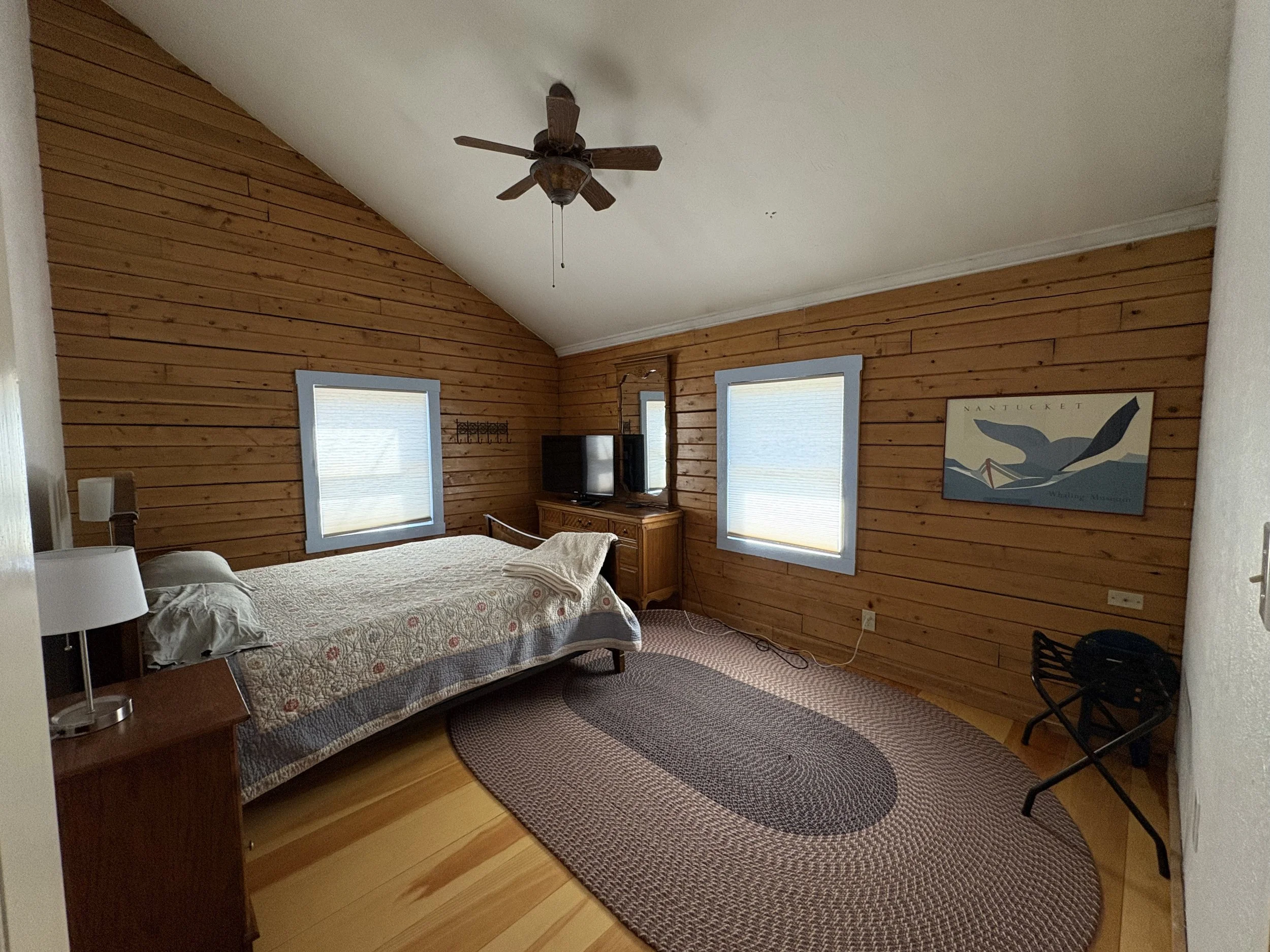 A bedroom with wooden paneled walls, a ceiling fan, two windows with blinds, a bed with a quilt, a small wooden nightstand with a lamp, a TV, and a framed picture of a whale on the wall.