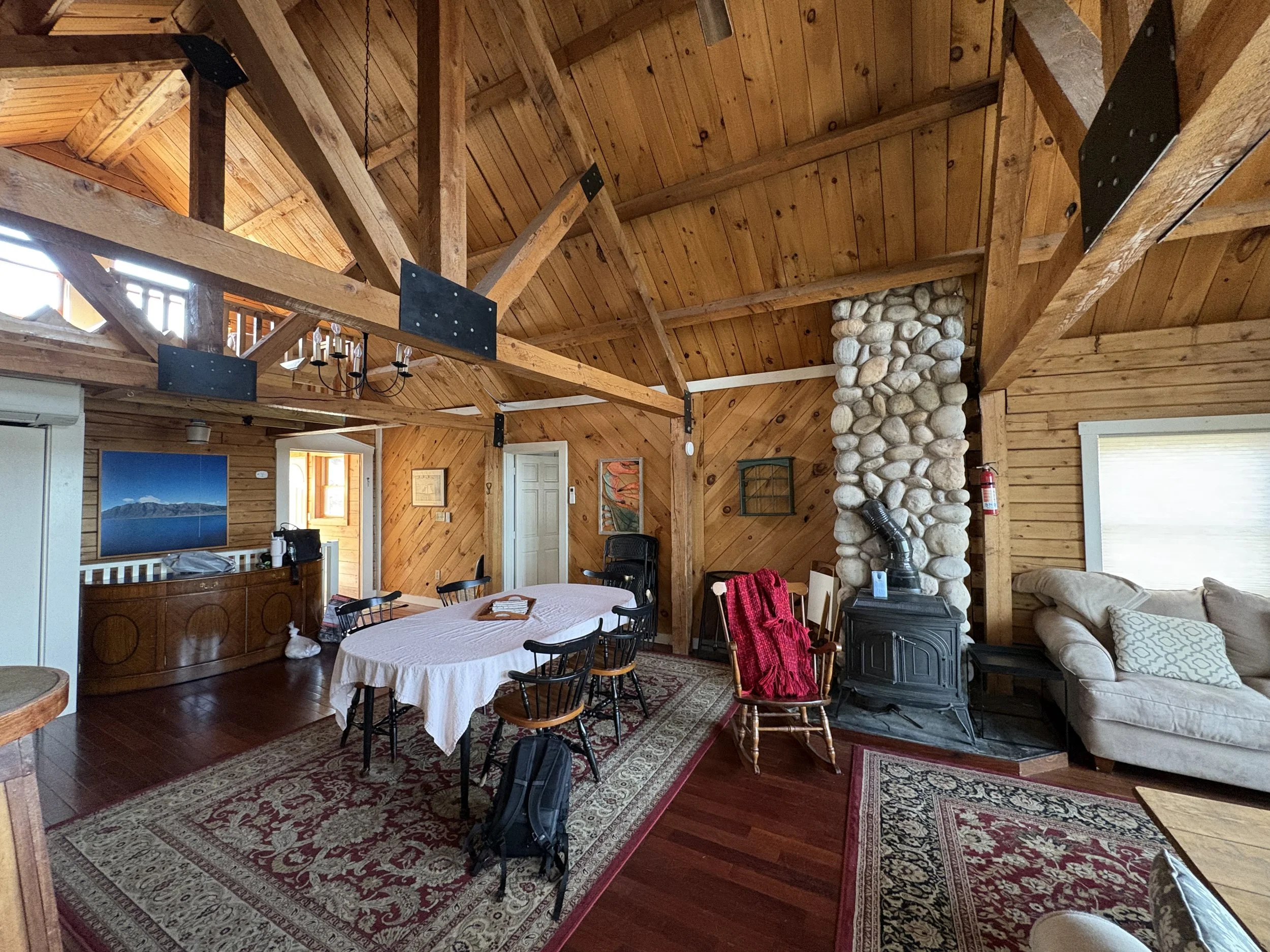 Interior view of a rustic wooden cabin living space with exposed wooden beams, a stone fireplace, a dining table with chairs, and a seating area with a sofa and patterned rugs.
