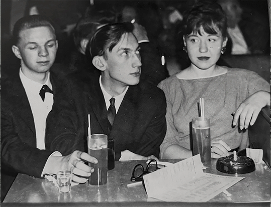 Three people sitting at a table with drinks and papers, engaged in conversation in a vintage black-and-white setting.